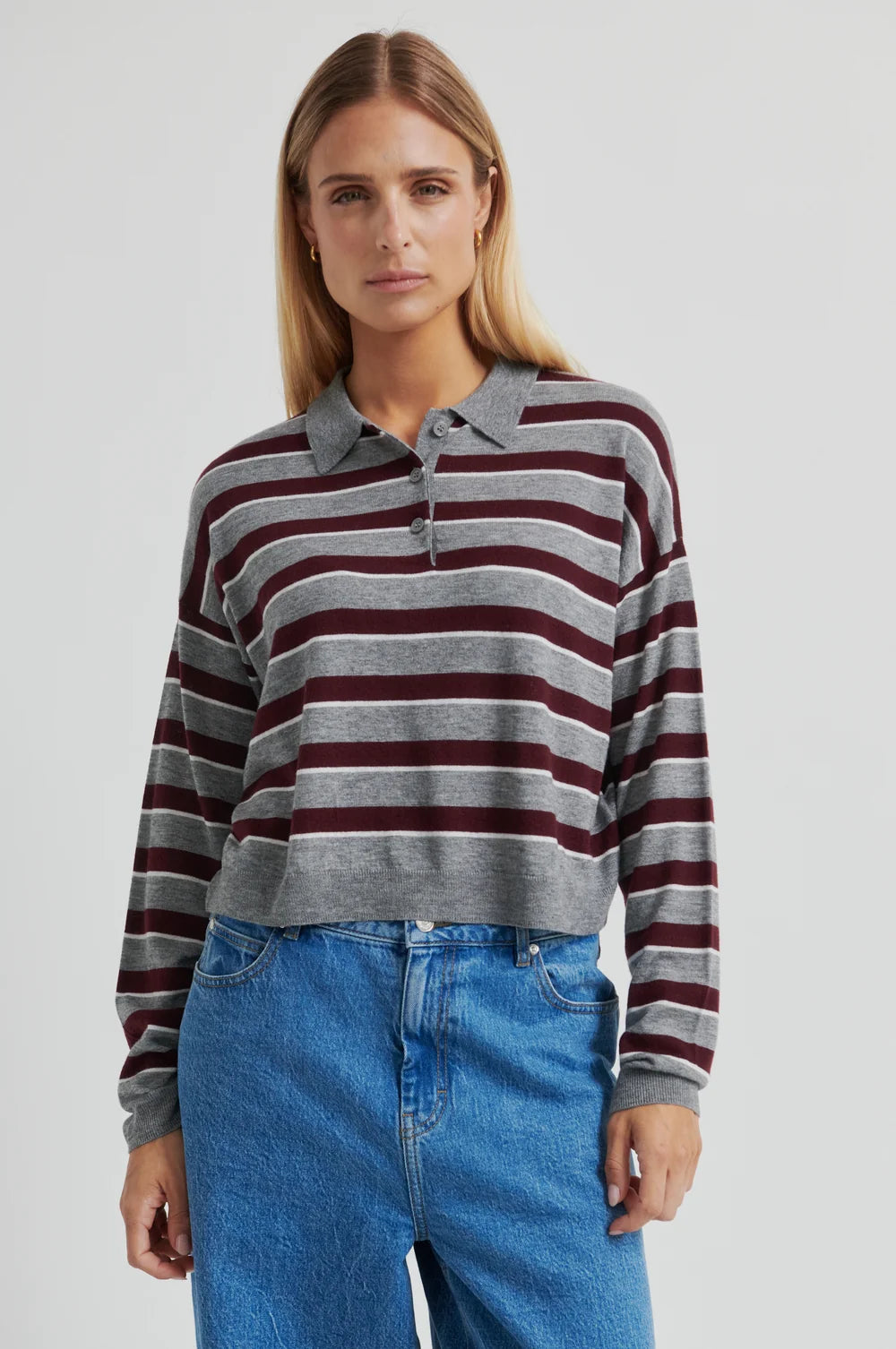 Women wearing a striped cropped polo sweater  in grey and burgundy with jeans on a plain background.  