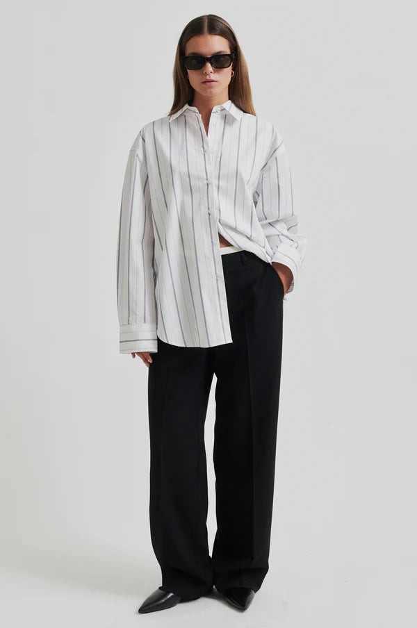 Model weasring white cotton shirt with black vertical stripes. Long sleeve and oversize fit.  Paired with black wide leg pants and black sunglasses. 
