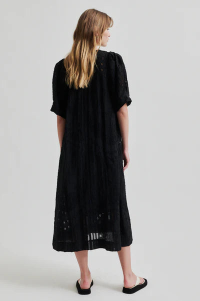 Intricate woven black midi dress with v neck and puff sleeves on model showing back of dress. 