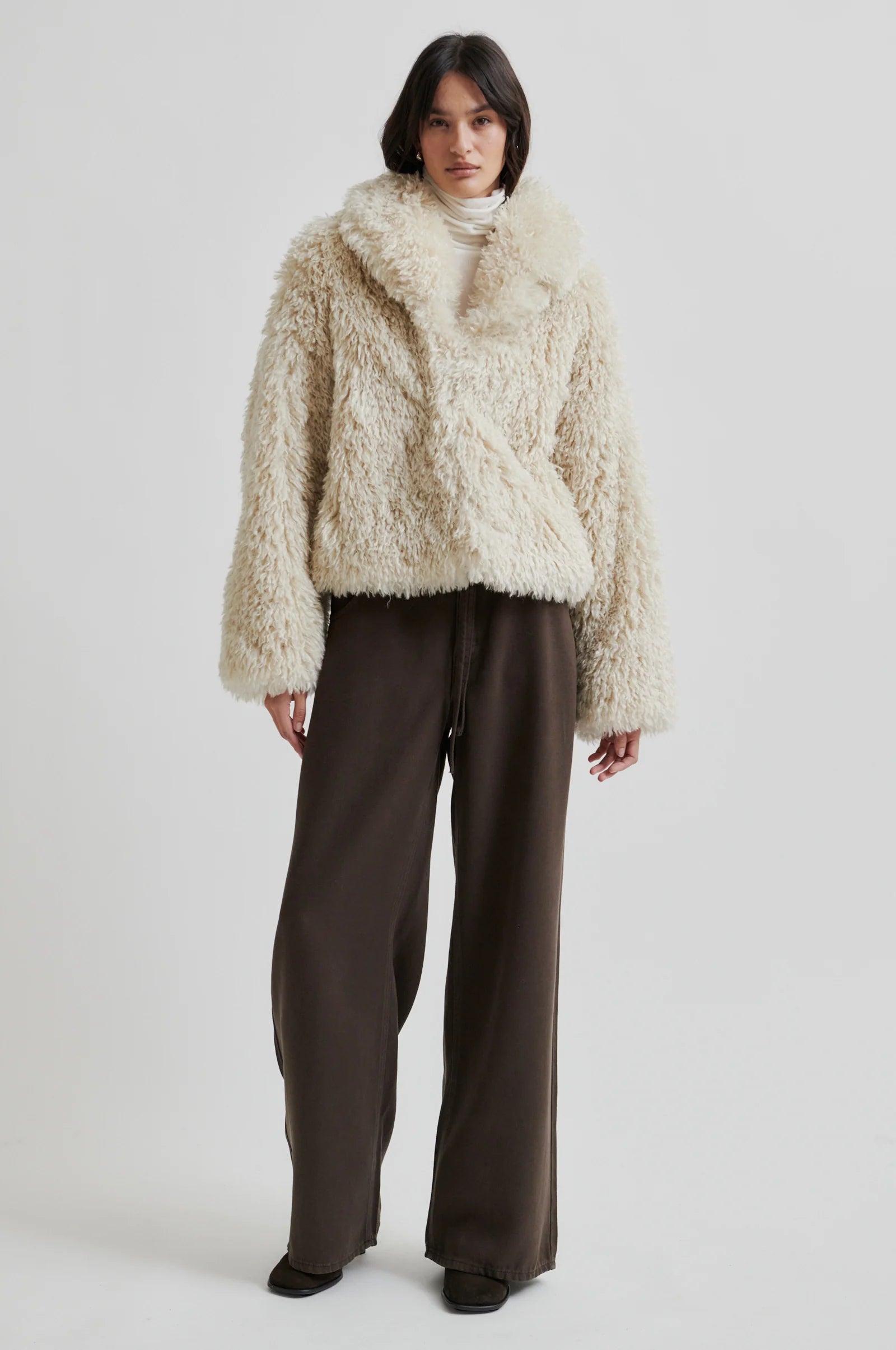 Person wearing a fluffy beige coat and brown pants on a white background