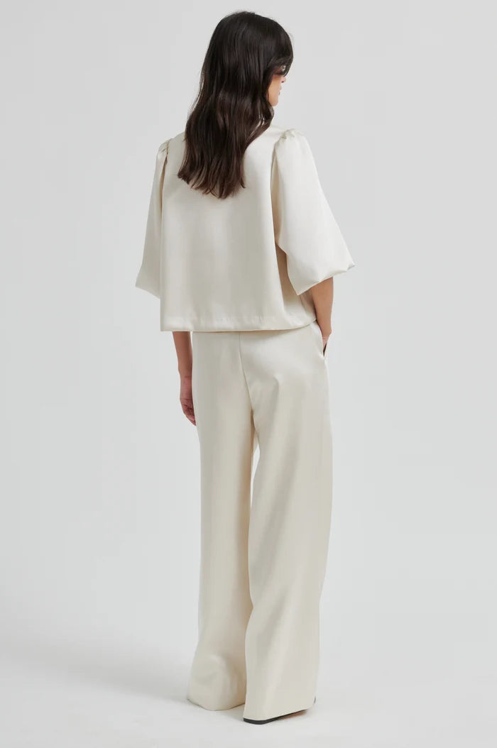 Woman from the back wearing a beige short sleeve blouse with ballon sleeves and matching pants on a plain background