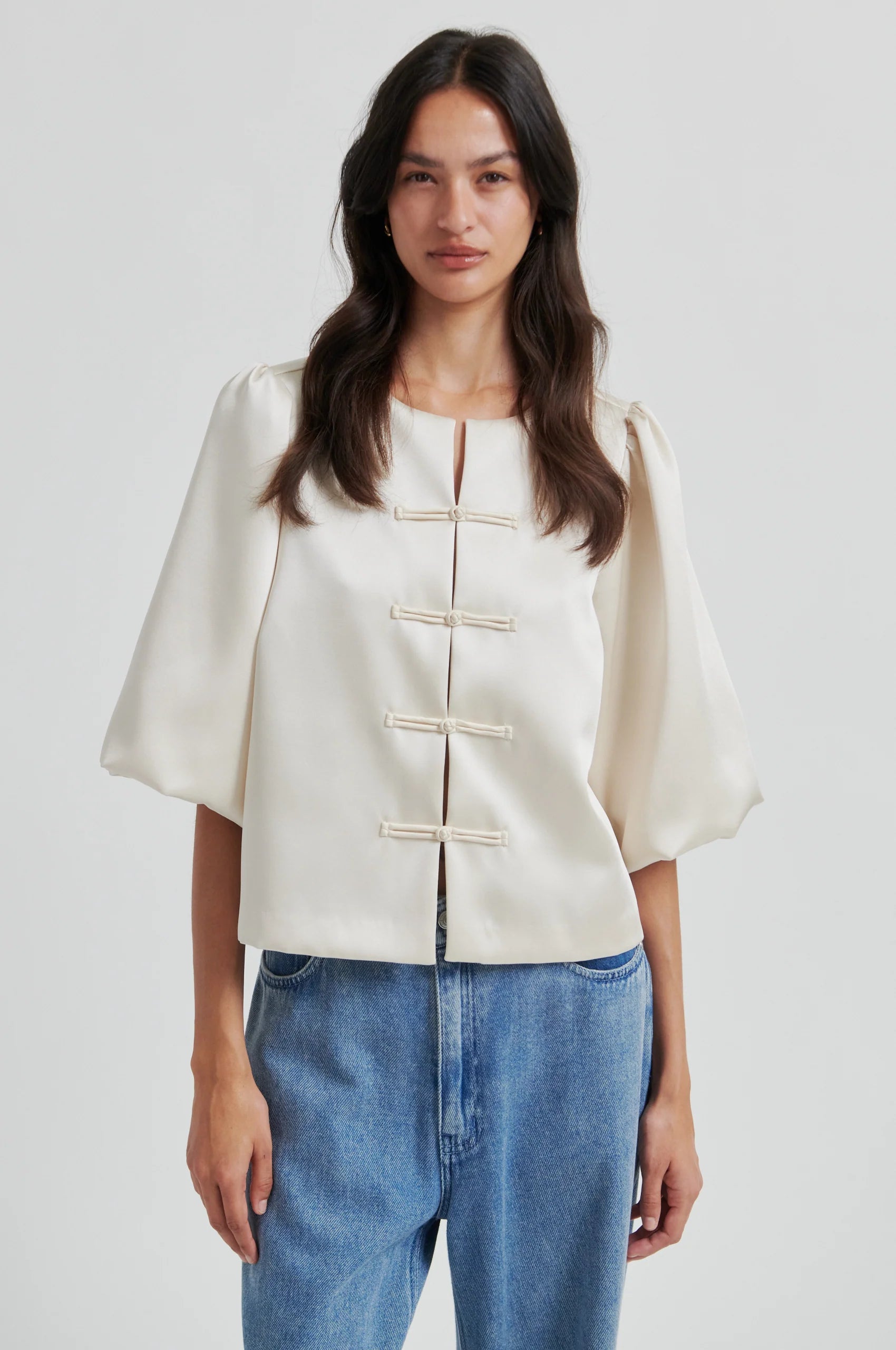 Woman wearing a beige short sleeve blouse with ballon sleeves and blue jeans on a plain background