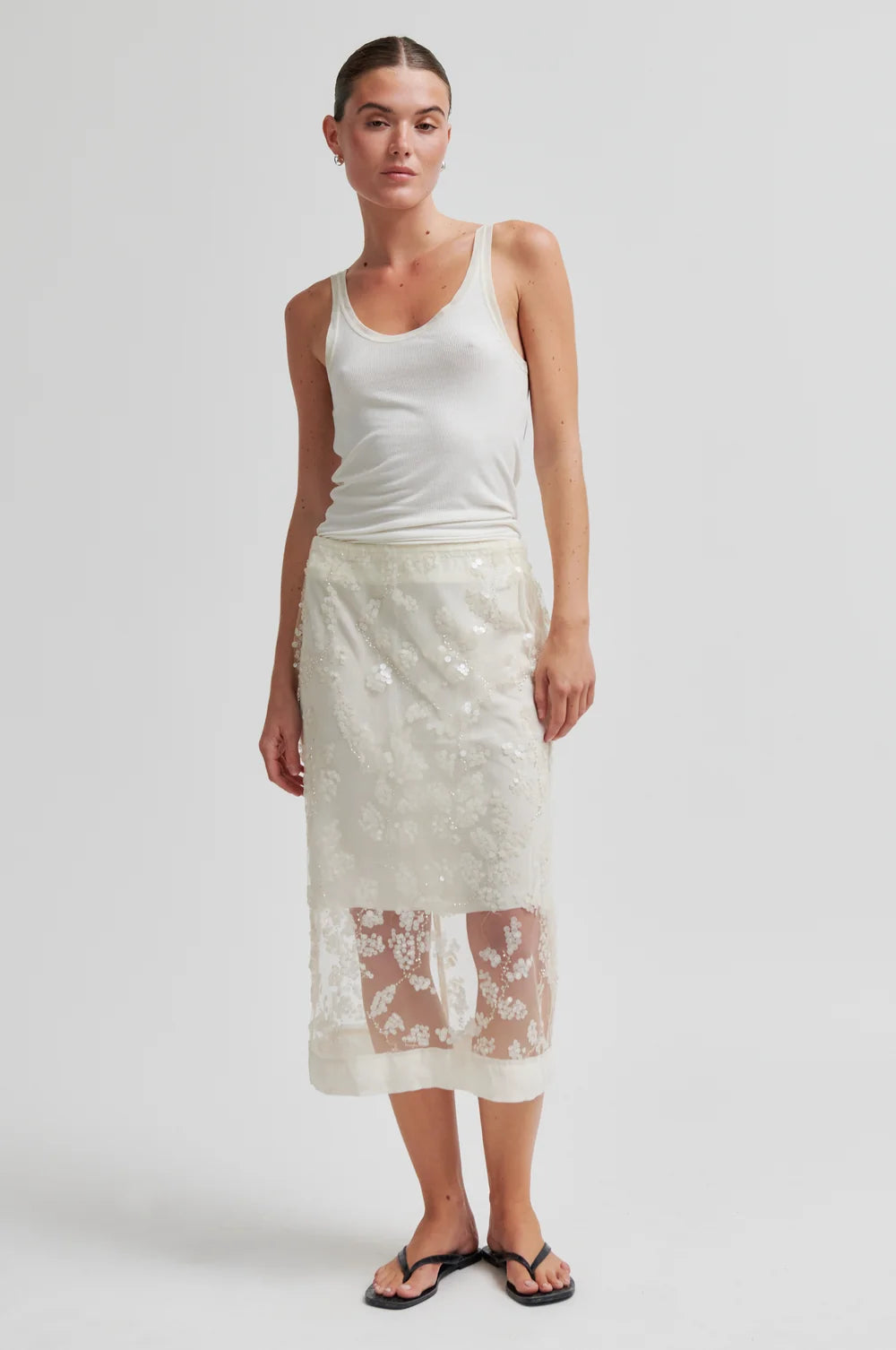 Woman wearing a white tank top and lace skirt on a plain background