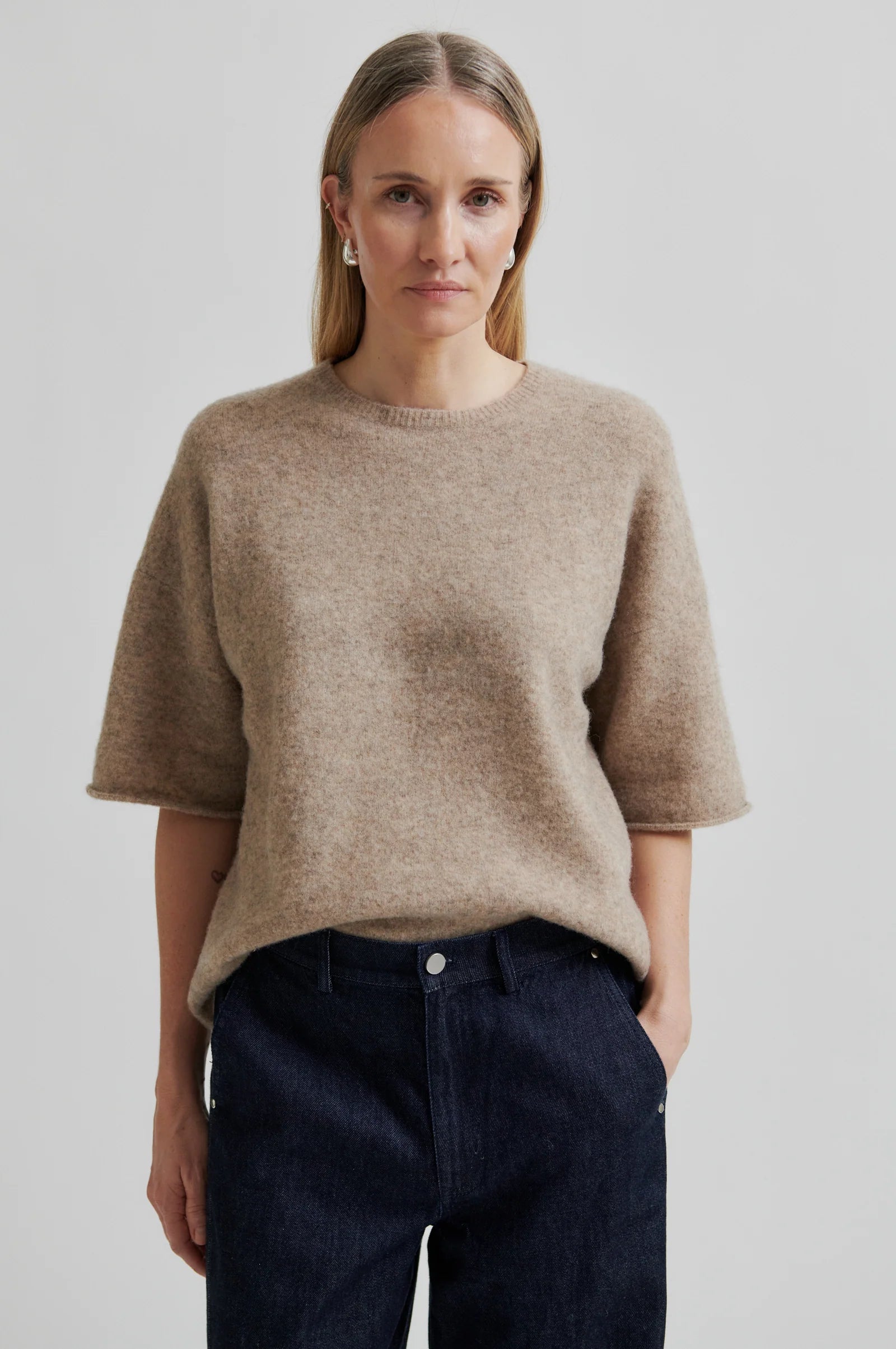 Woman wearing a beige short sleeve sweater and dark jeans against a plain background