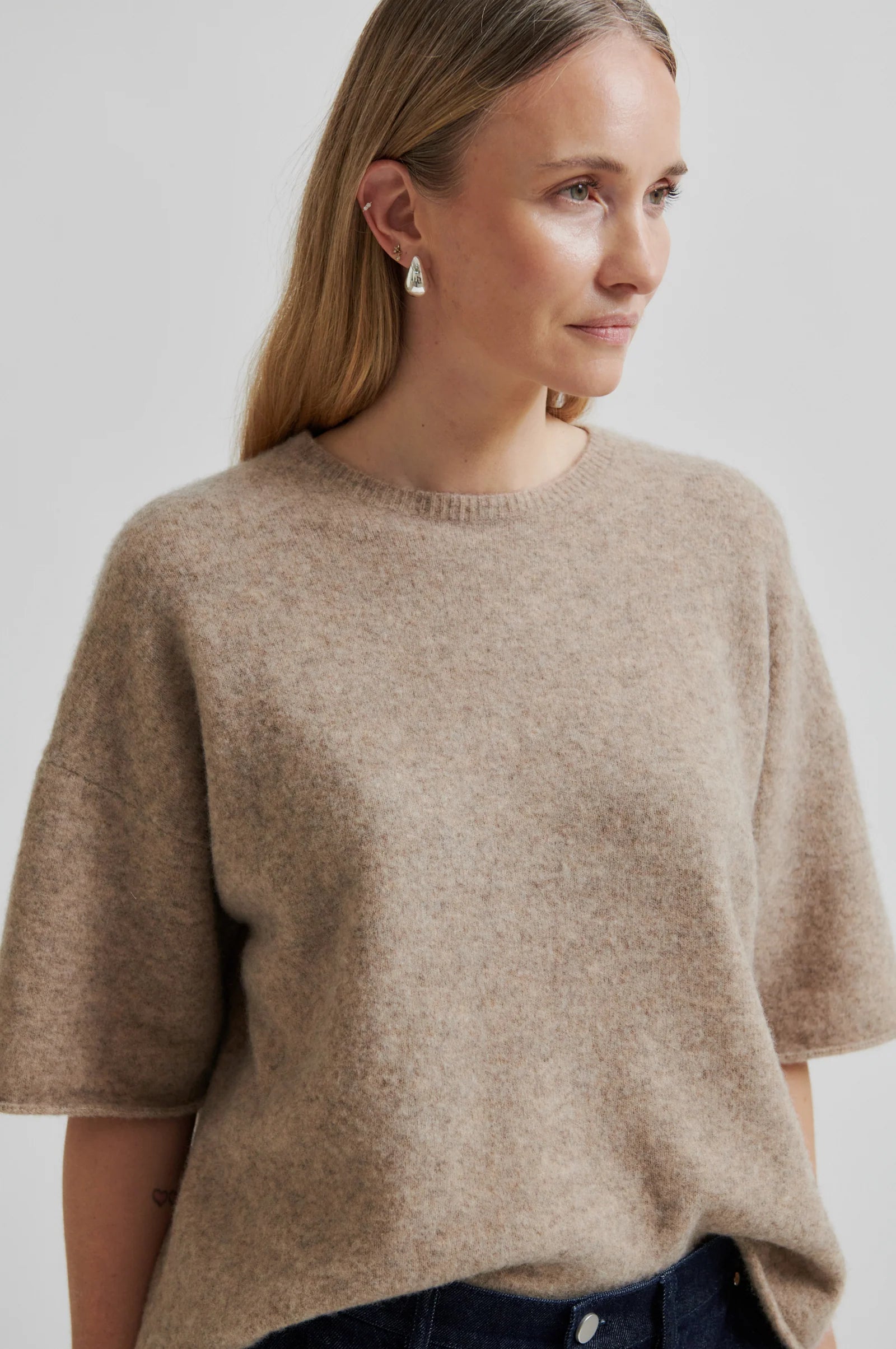 Woman wearing a beige short sleeve sweater and dark jeans against a plain background