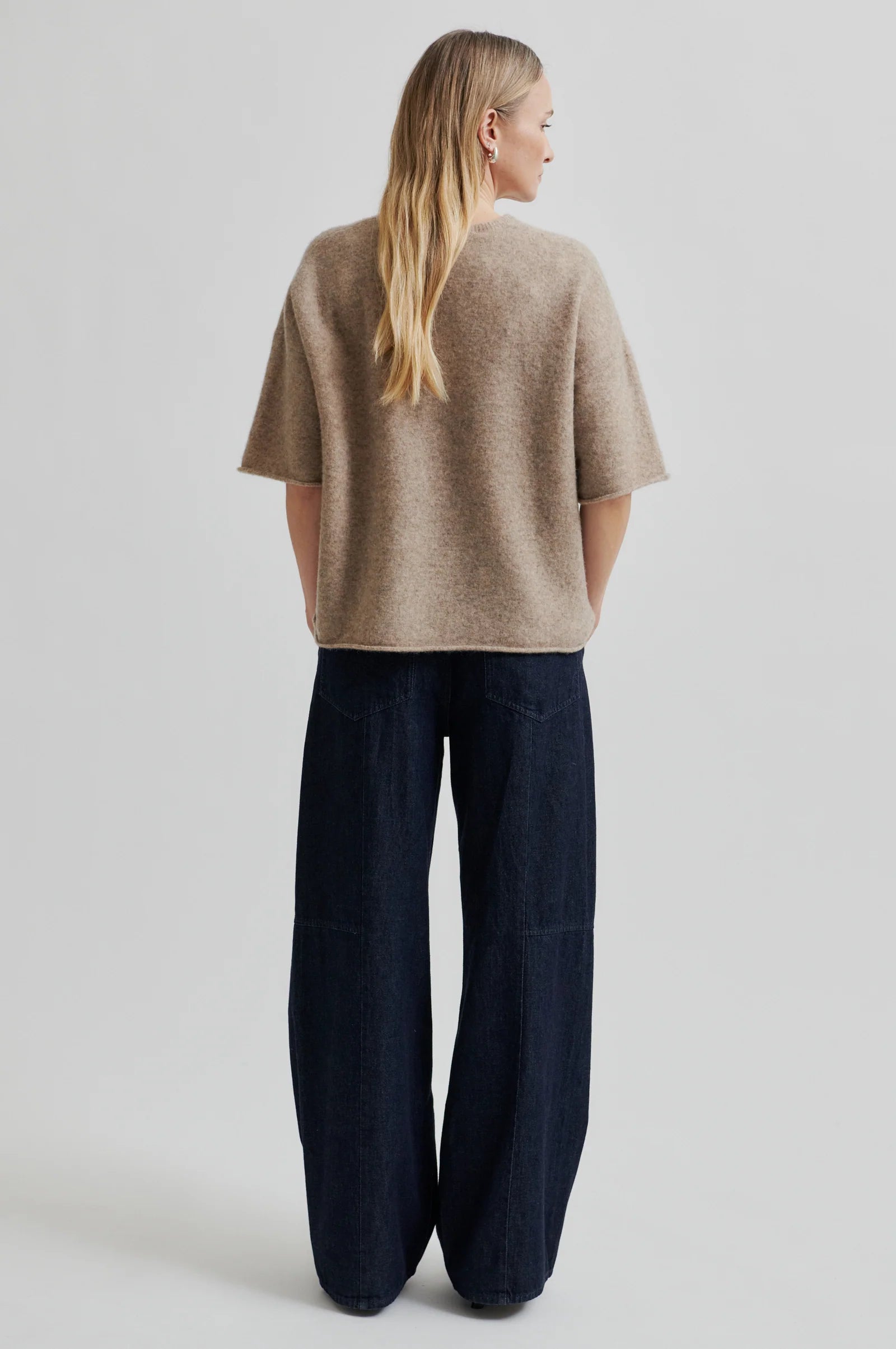 Woman wearing a beige short sleeve sweater and dark jeans against a plain background