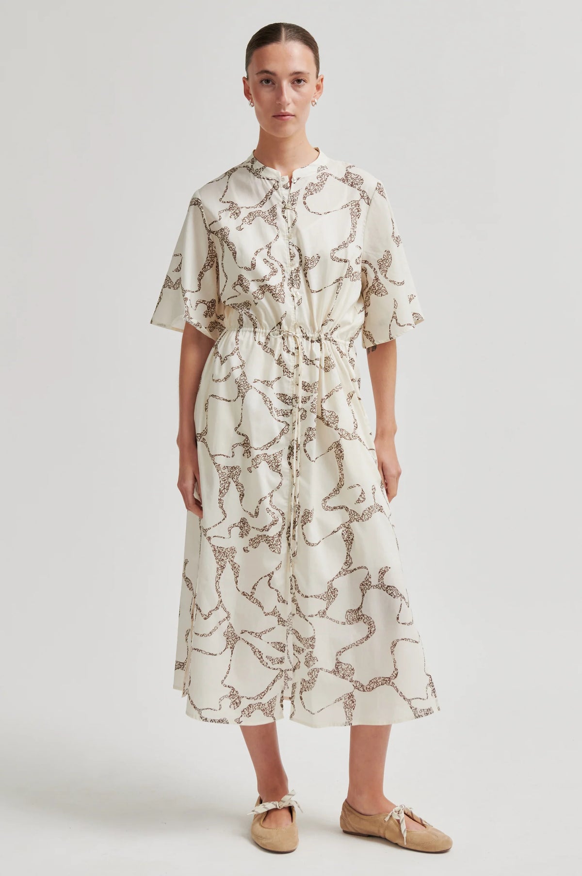 A woman stands against a light gray studio background wearing a short-sleeve midi dress with an abstract chain-link pattern in beige and brown on an off-white base. She is also wearing tan flat shoes.