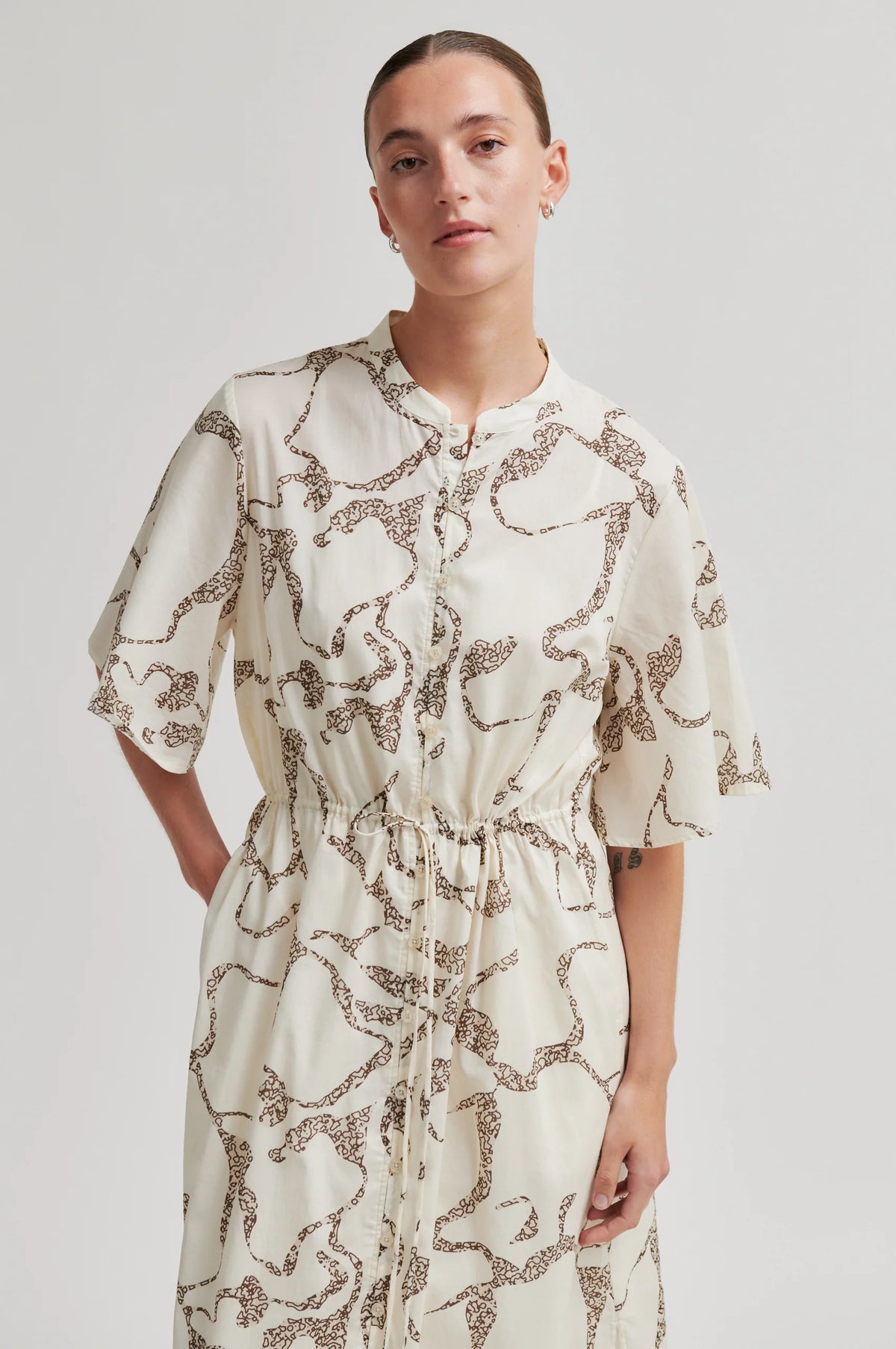A woman wearing a cream-colored short-sleeve dress with a brown abstract pattern, standing against a light gray studio background.