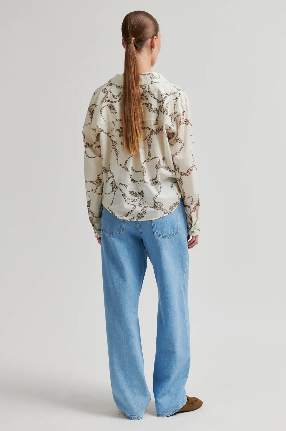 Woman shown from the back with long ponytail wearing a patterned shirt and blue jeans on a plain background
