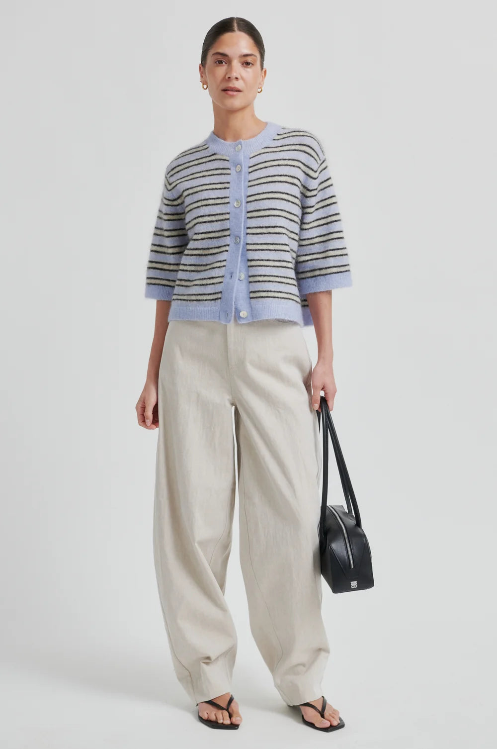 Woman wearing a blue cream and brown striped short sleeve cardigan and wide-leg pants with a black handbag on a light background