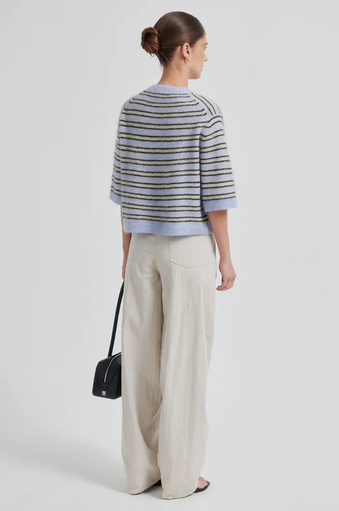 Woman wearing a blue cream and brown striped short sleeve cardigan and wide-leg pants with a black handbag on a light background