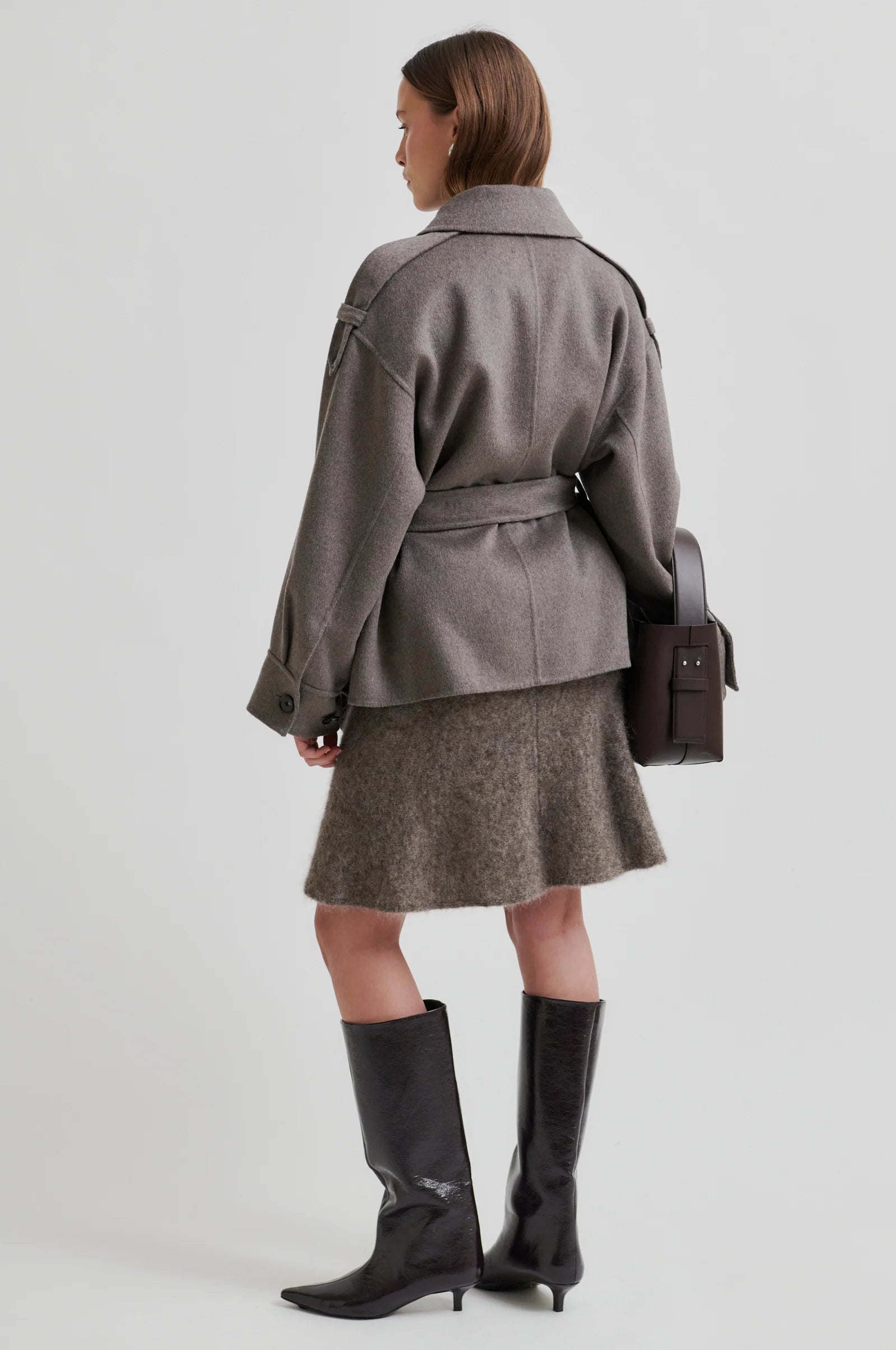 Woman wearing a light brown coat with belt, skirt, and black boots on a white background