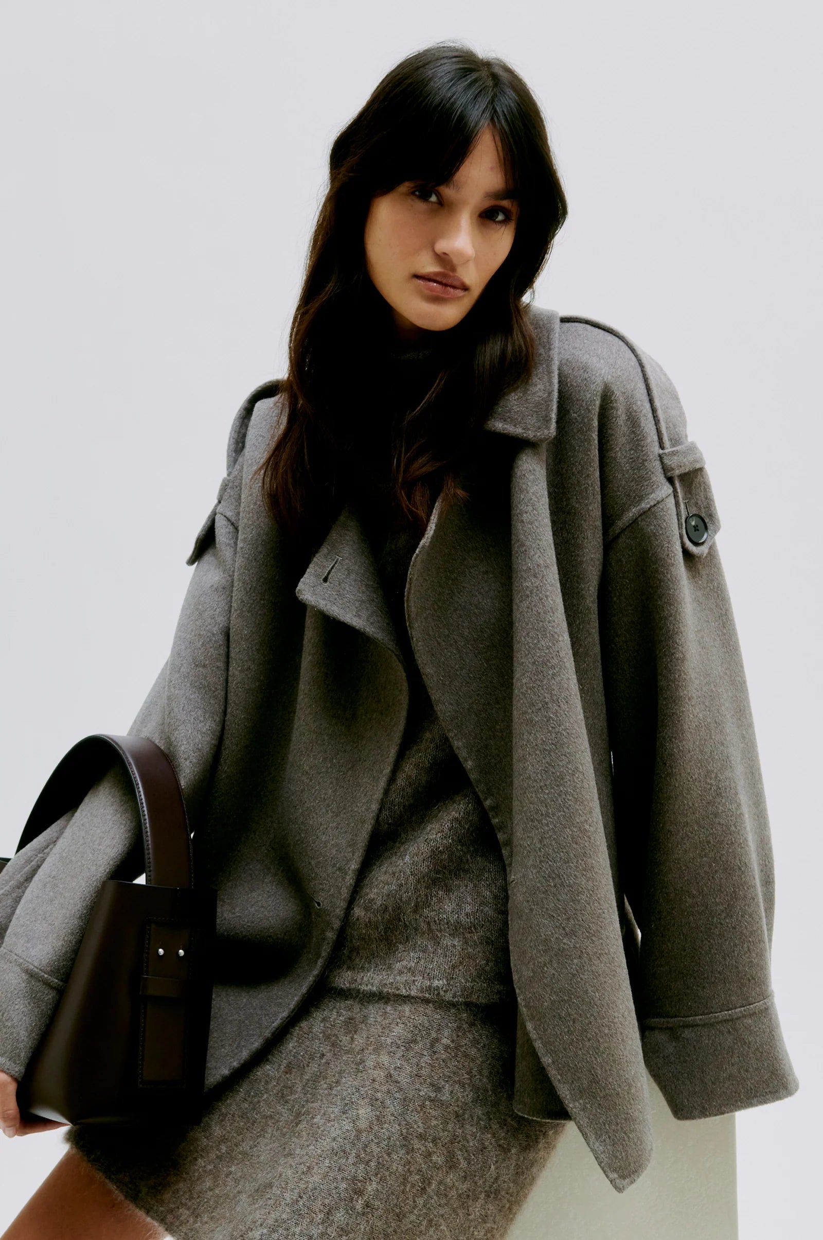 Woman wearing a light brown coat and skirt and dark brown bag set against a plain background