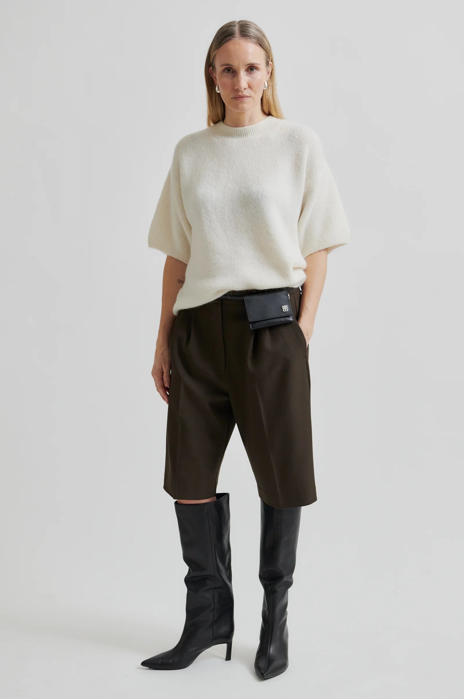 Woman wearing a short sleeve white sweater and dark pants with a black bag on a plain background