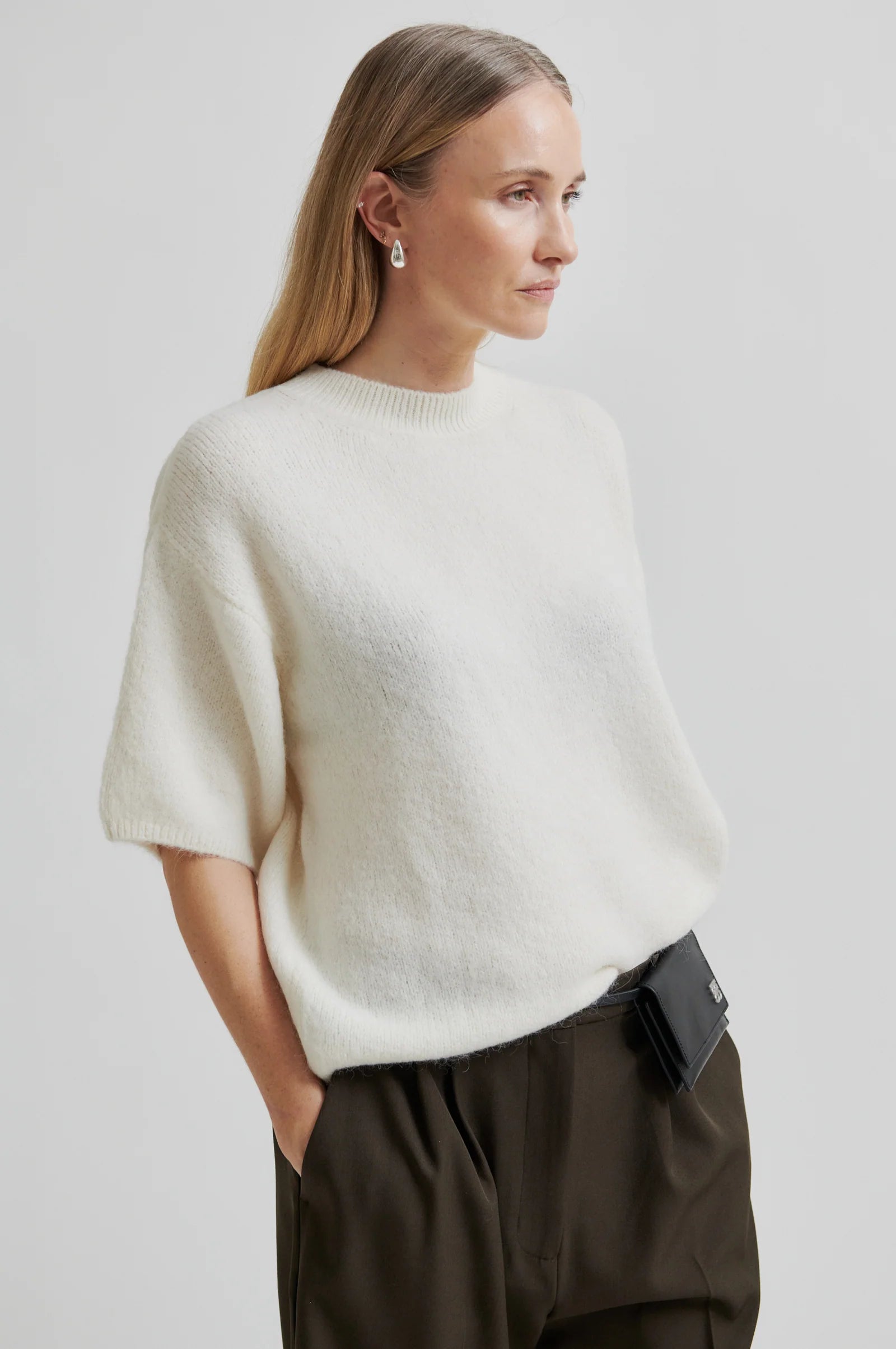 Woman wearing a short sleeve white sweater and dark pants with a black bag on a plain background up close 