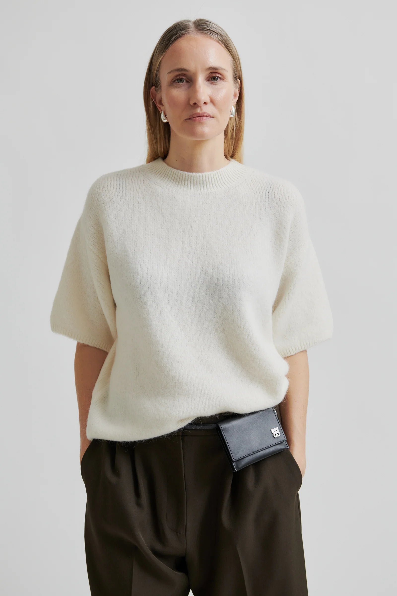 Woman wearing a short sleeve white sweater and dark pants with a black bag on a plain background