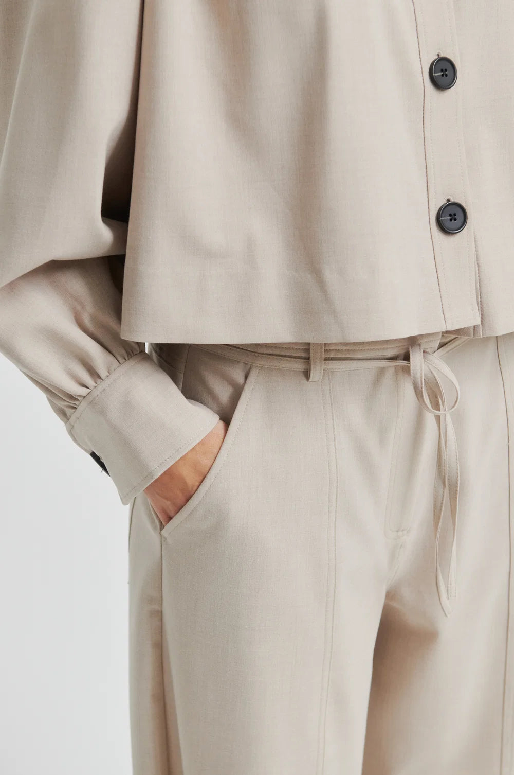 Close up of lady wearing beige wide-leg pants and top in same colour with black buttons on a plain background