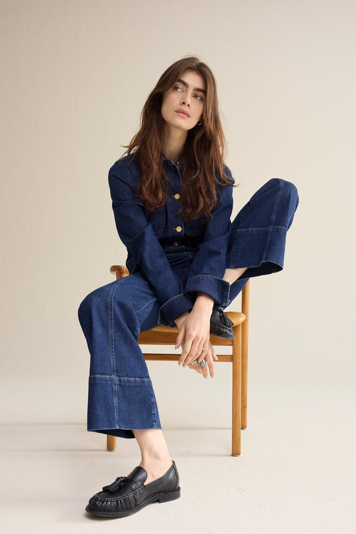 Woman sitting on a chair posing wearing a blue denim jacket and cropped jeans with slight flare  on a white background