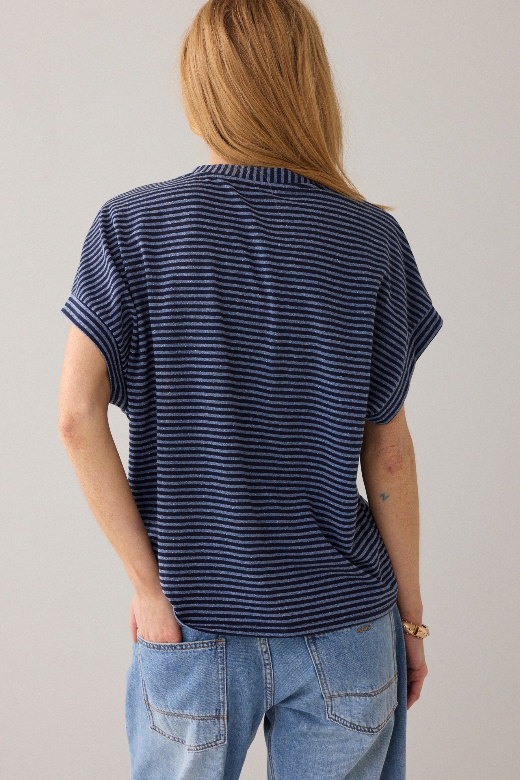Women shown from the back short-sleeve crewneck knit top with horizontal navy and light blue stripes and light blue jeans on a solid light beige background.