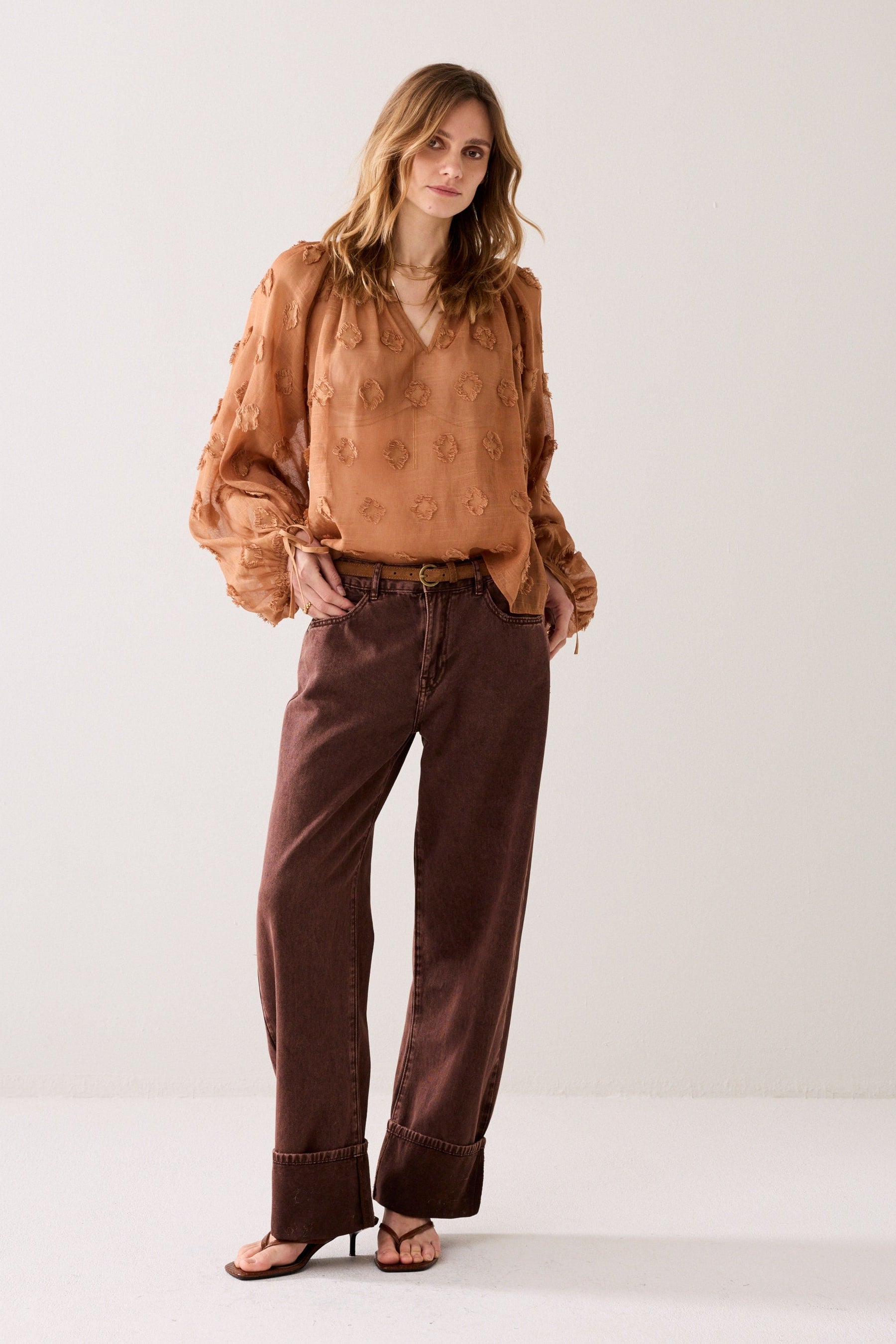 Woman wearing a cinnamon embroidered blouse with gold jewellery and burgundy jeans on a plain background.