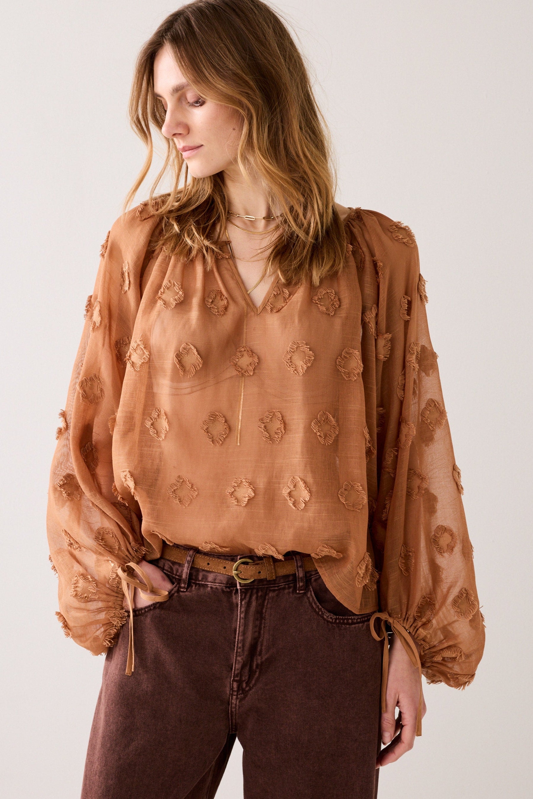 Woman wearing a cinnamon embroidered blouse with gold jewellery on a plain background.