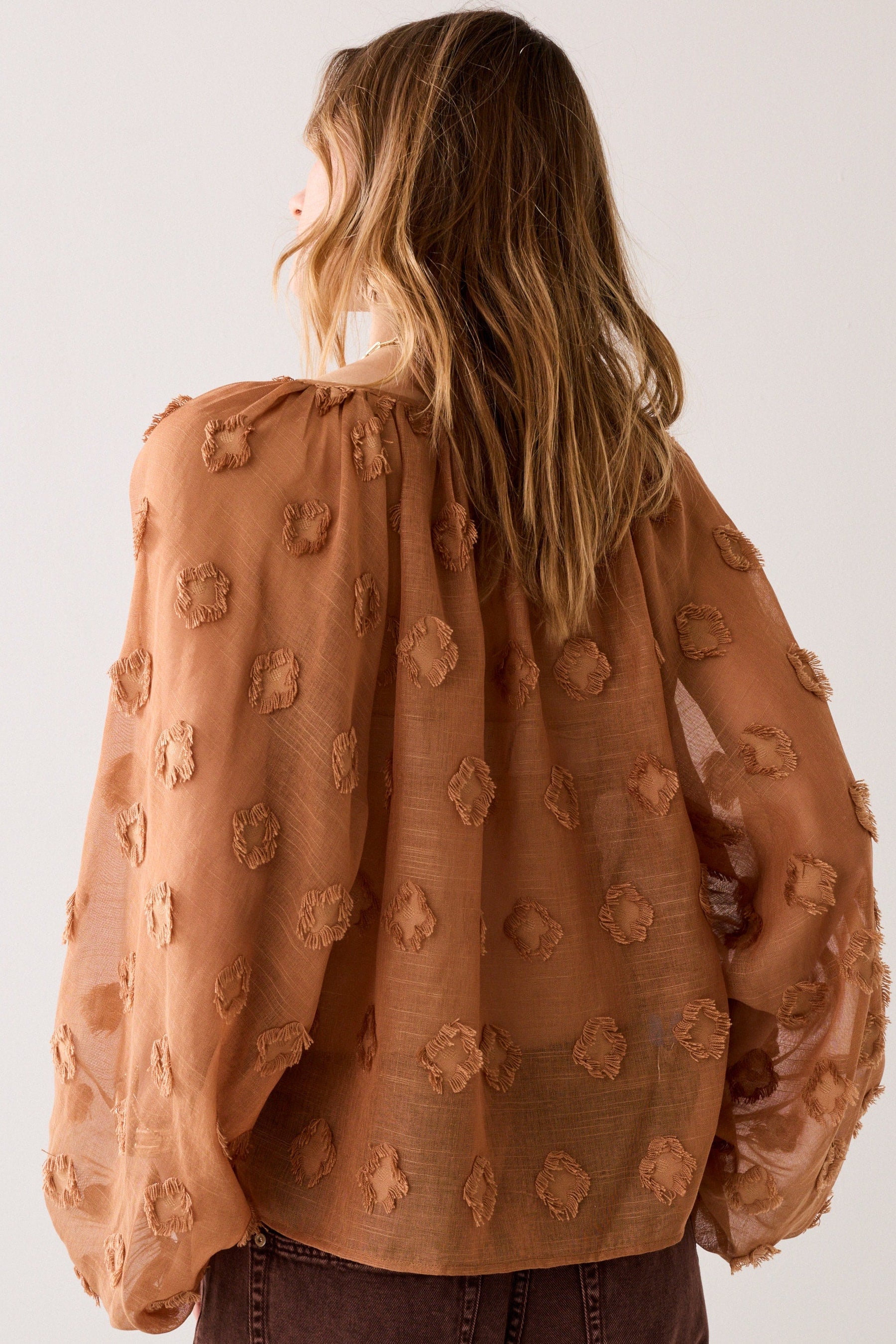 Woman shown from the back wearing a cinnamon embroidered blouseon a plain background.