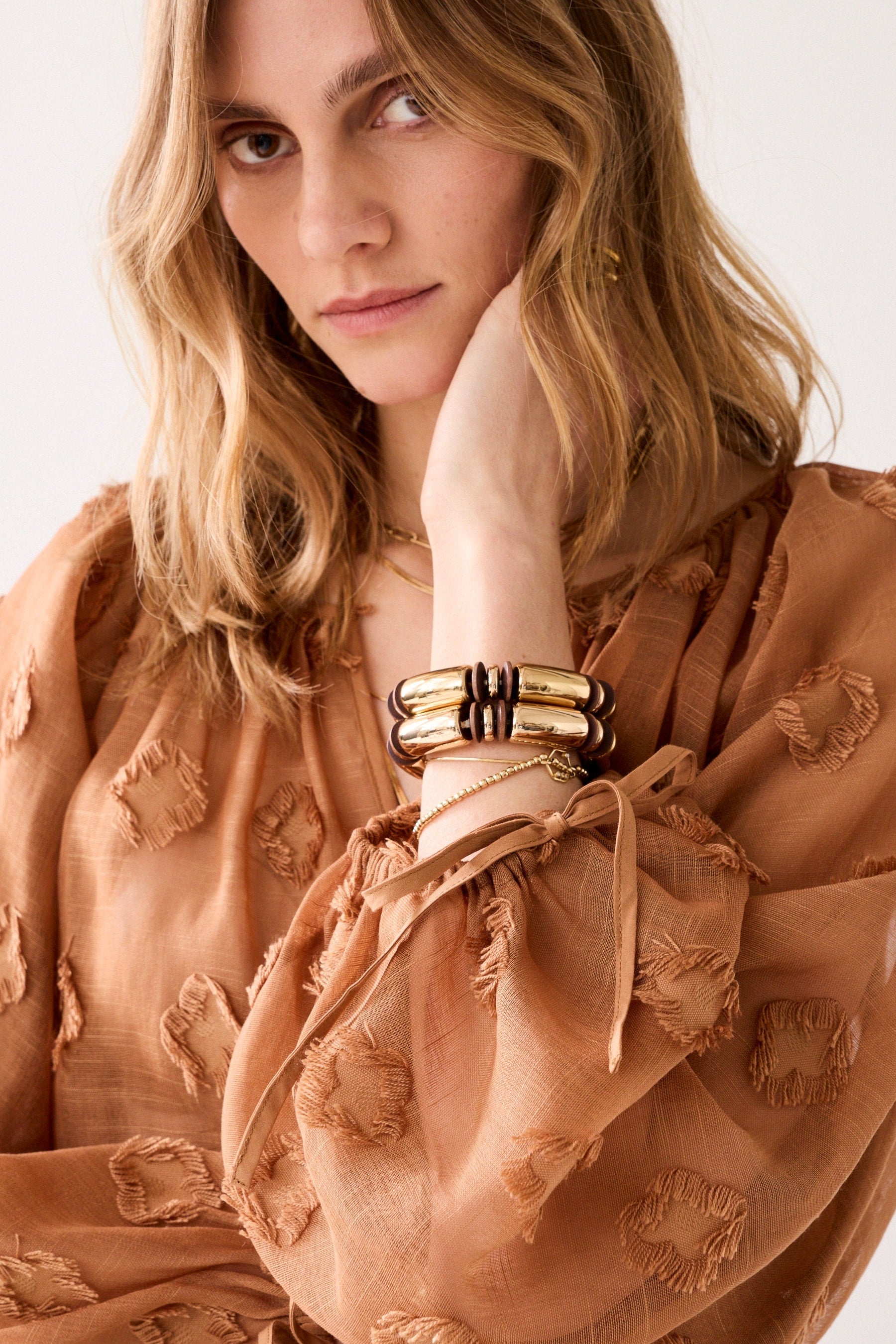Woman wearing a cinnamon embroidered blouse with gold jewellery on a plain background.