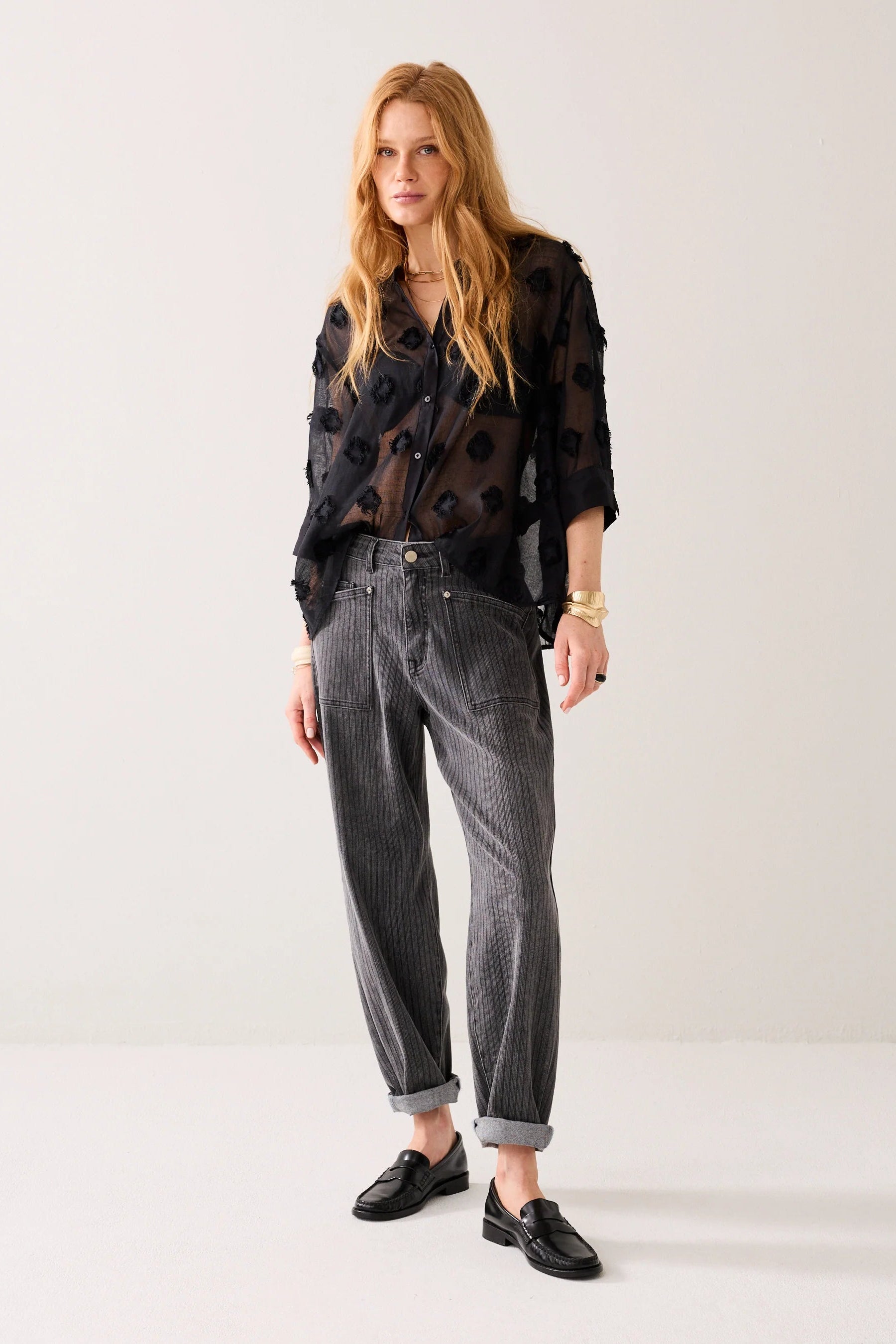 Woman wearing a black sheer blouse with a 3d  pattern with light grey stripe jeans, black loafers and gold jewellery on a plain background