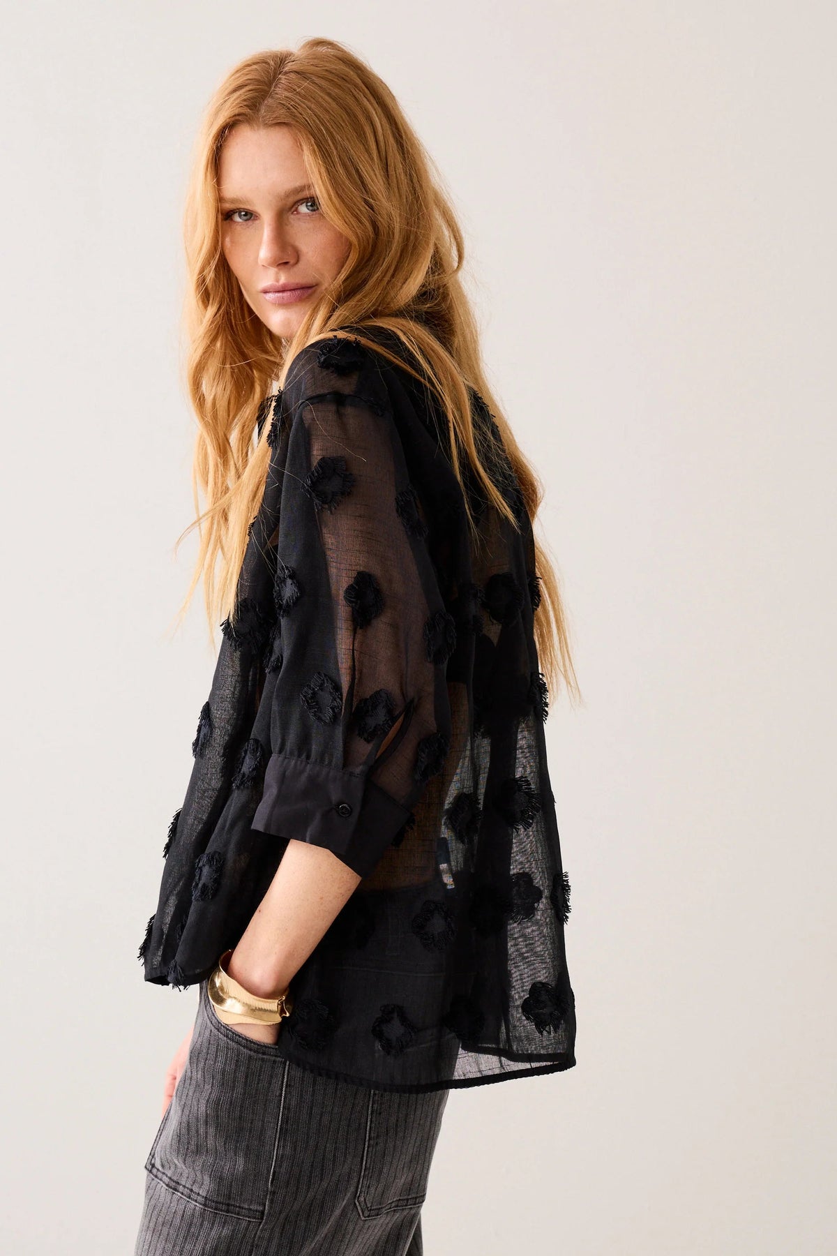 Woman shown side on wearing a black sheer blouse with a 3d  pattern with light grey stripe jeans and gold jewellery on a plain background
