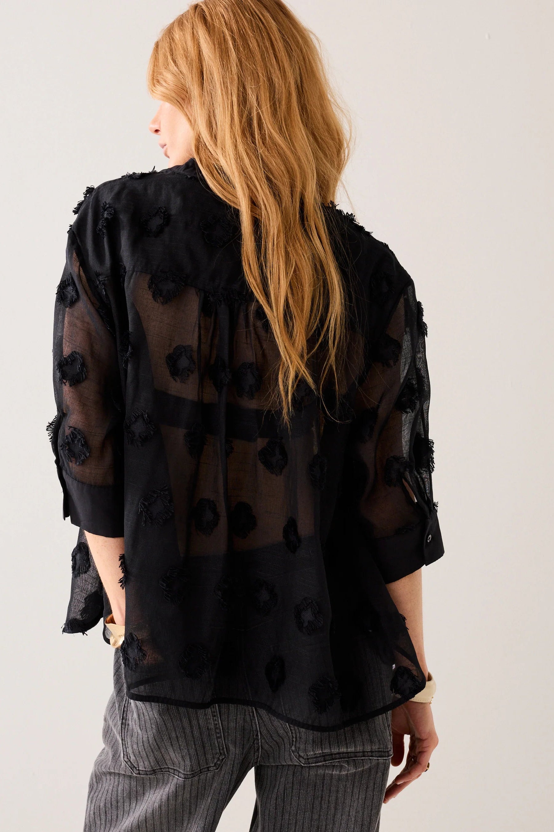 Woman shown from the back wearing a black sheer blouse with a 3d  pattern with light grey stripe jeans on a plain background