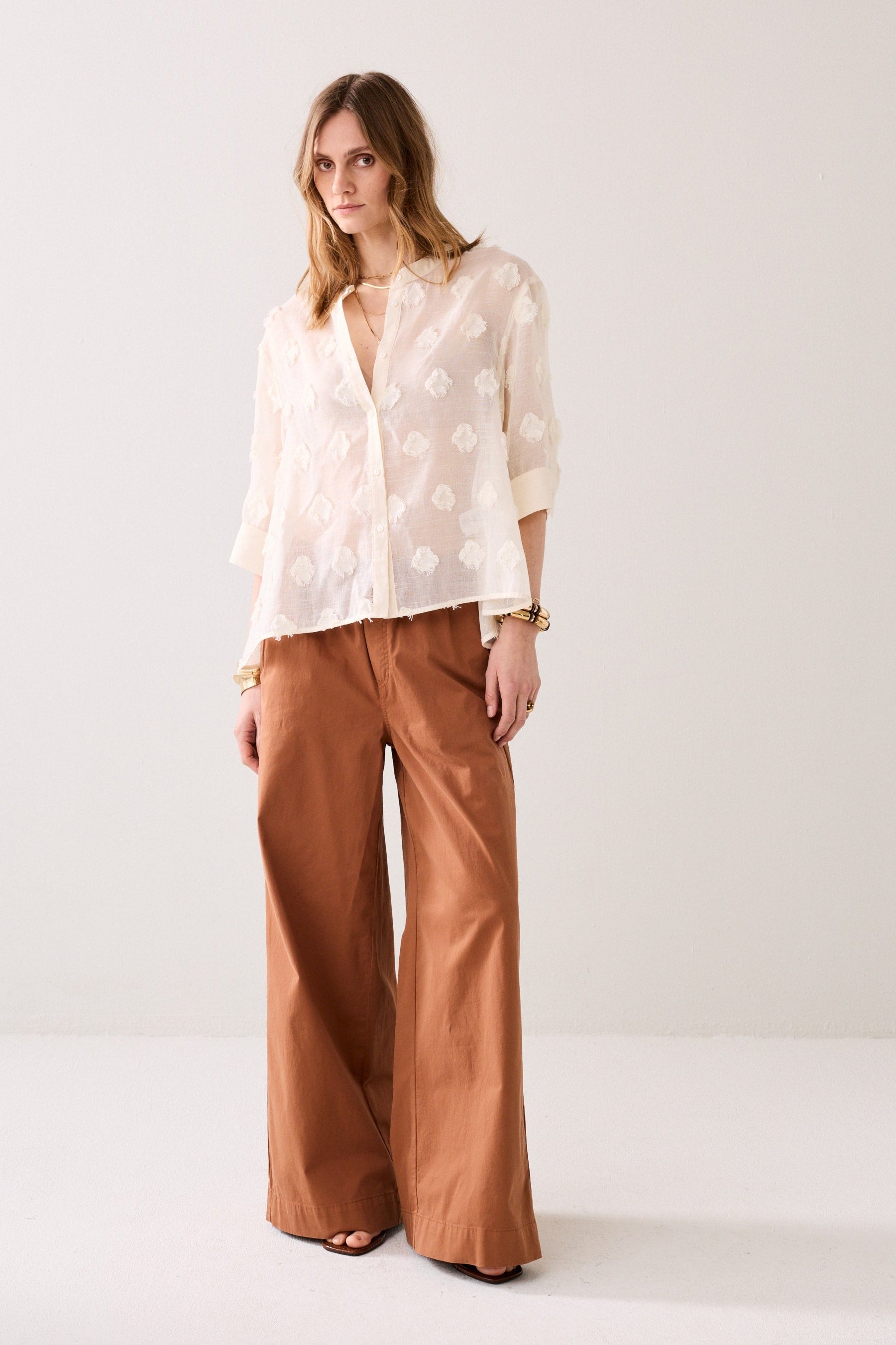 Woman wearing a ivory coloured blouse with 3d floral patterns and brown pants with gold jewellery against a white background