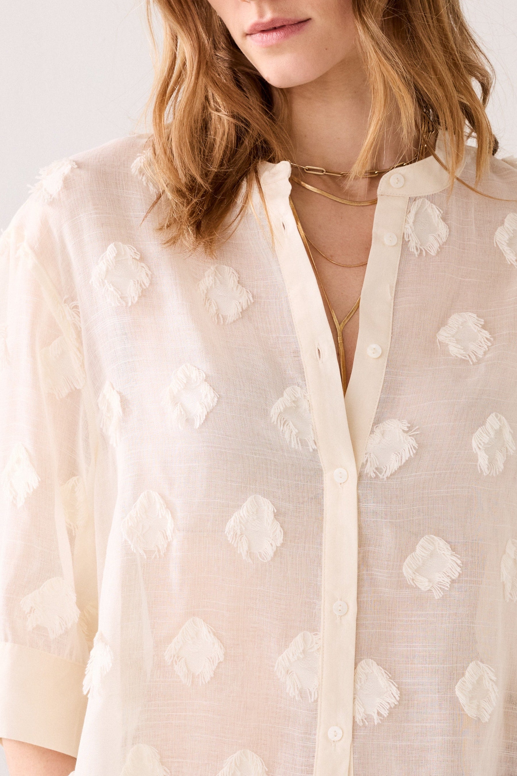 Woman up close wearing a ivory coloured blouse with 3d floral patterns and gold necklaces against a white background