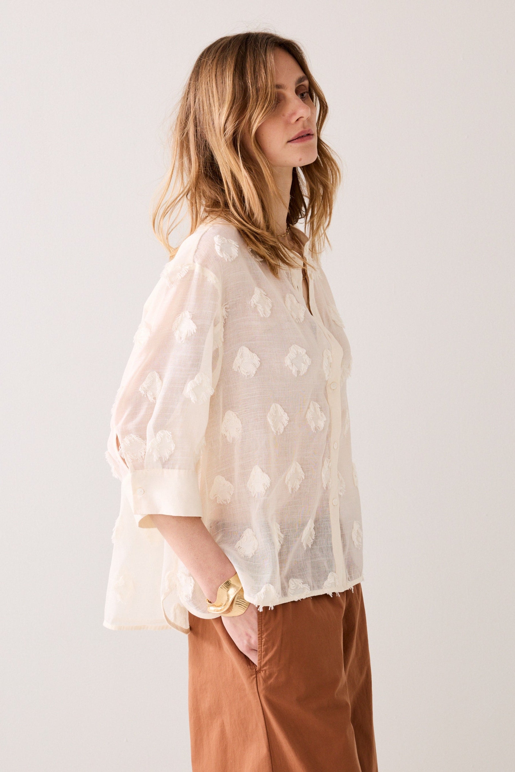 Woman wearing a ivory coloured blouse with 3d floral patterns and brown pants against a white background
