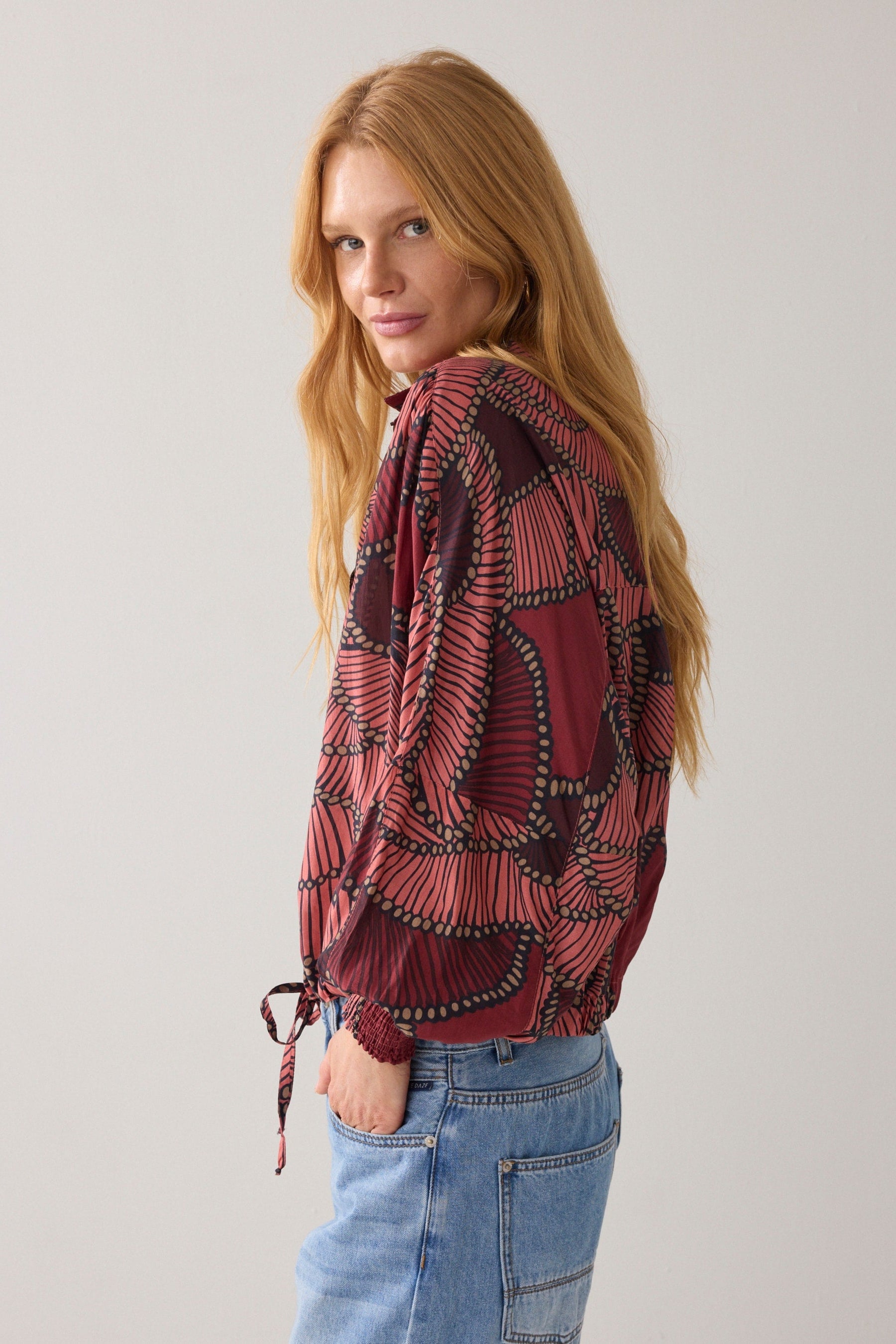 Woman wearing a patterned blouse and jeans against a plain background