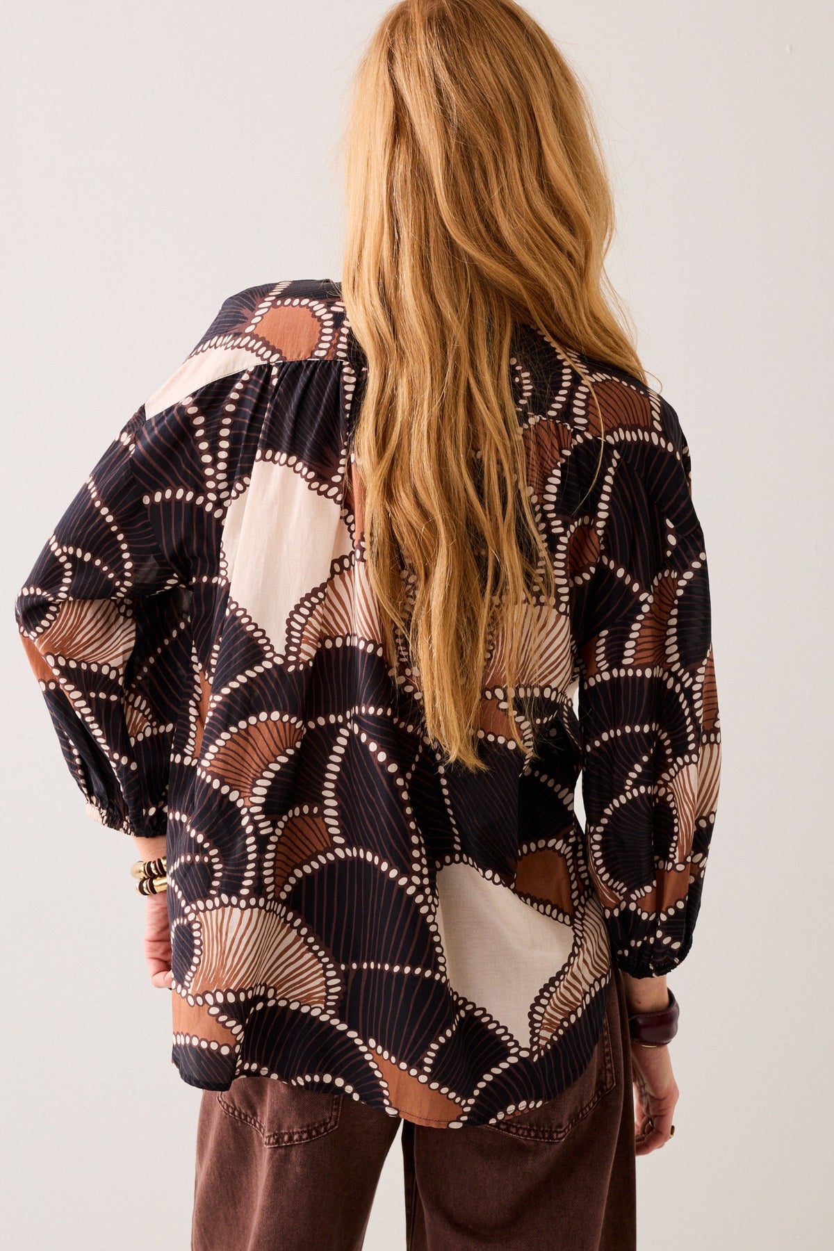 A women shown from the back wearing a multi colour long-sleeve patterned shirt with brown pants on plain background