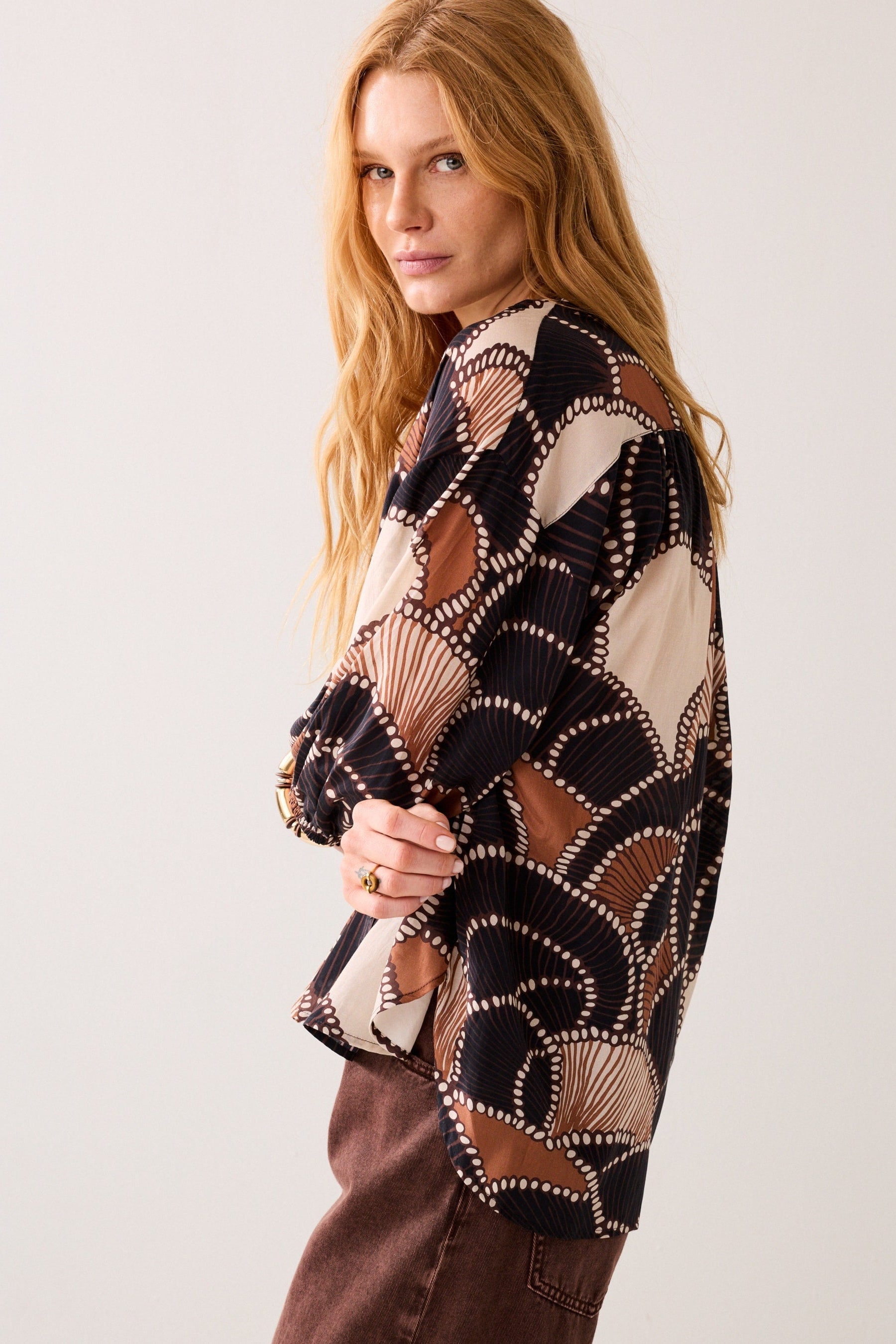 A women side on wearing a multi colour long-sleeve patterned shirt with brown pants on plain background