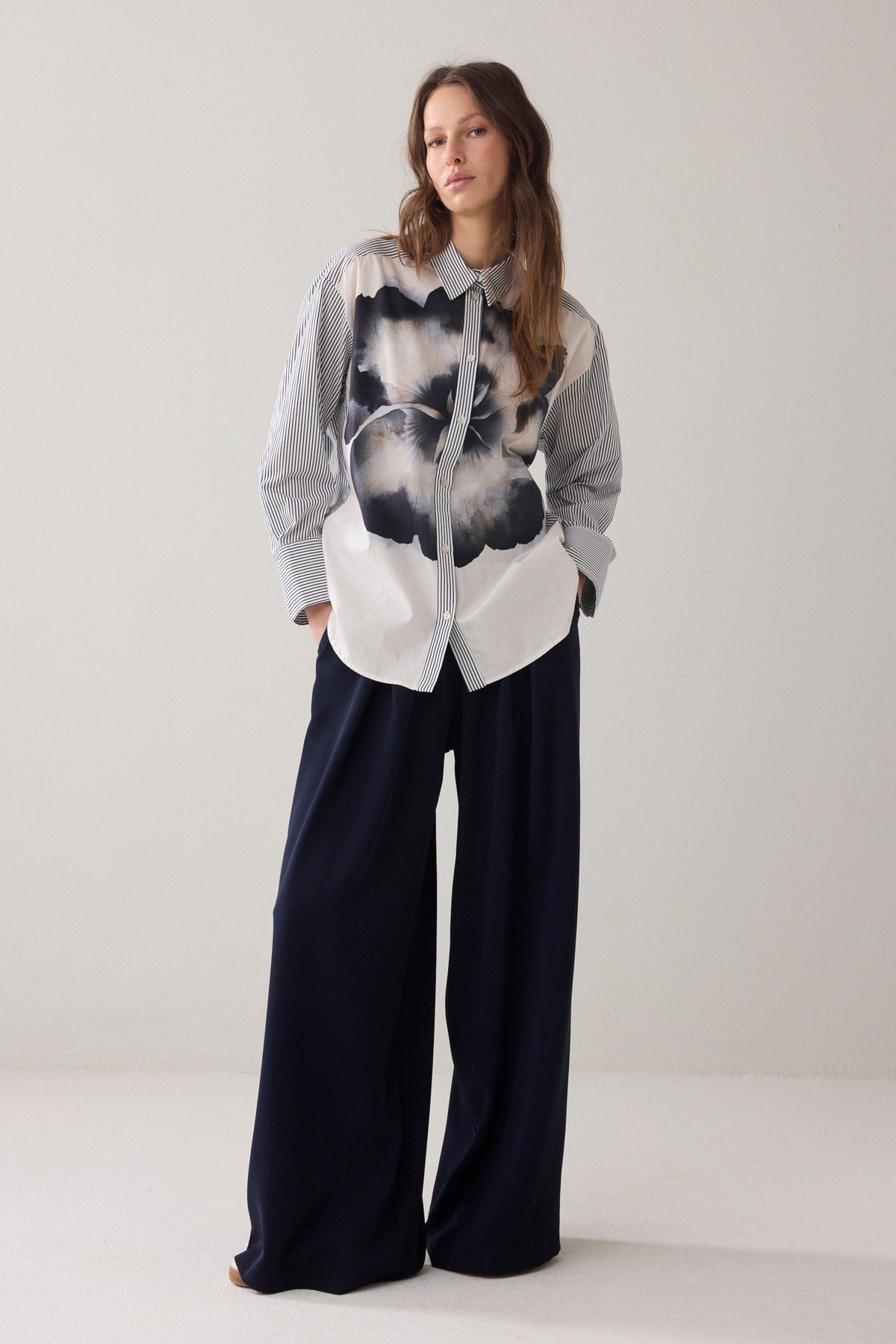 A woman  wearing a long-sleeve button-up shirt with thin vertical blue and white stripes, and statement floral design paired with dark pants, against a plain light beige background.

