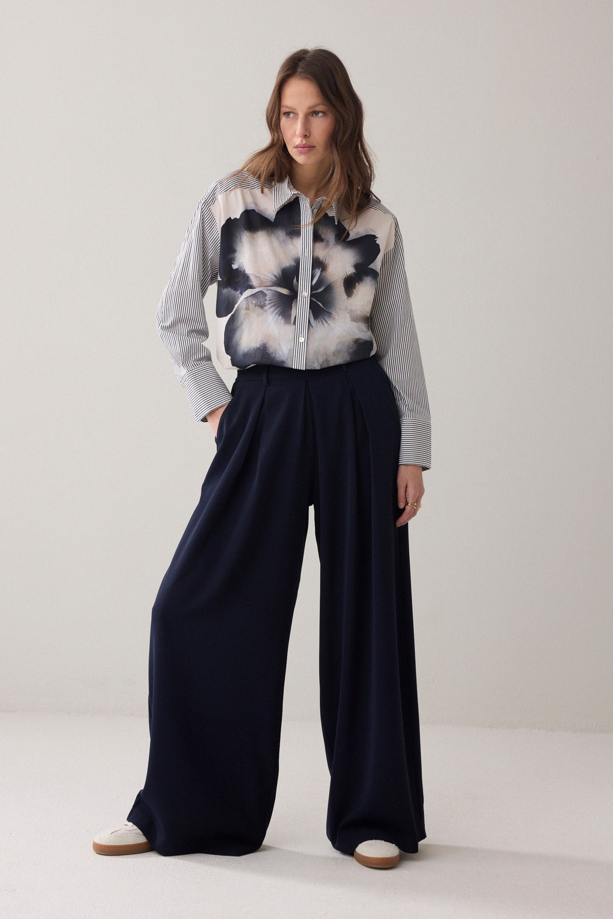 A woman  wearing a long-sleeve button-up shirt with thin vertical blue and white stripes, and statement floral design paired with dark pants, against a plain light beige background.