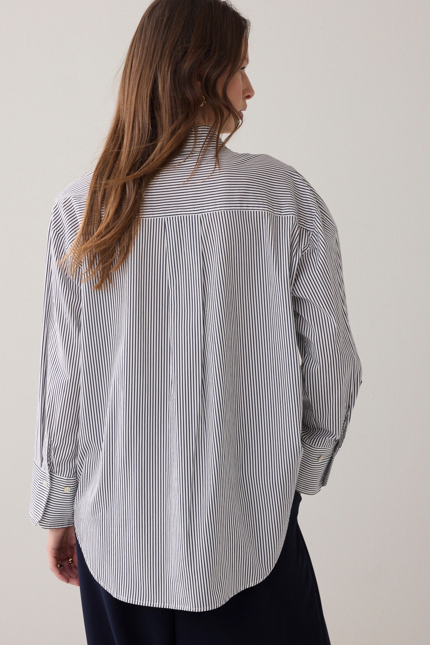 A woman shown from the back wearing a long-sleeve button-up shirt with thin vertical blue and white stripes, paired with dark pants, against a plain light beige background.