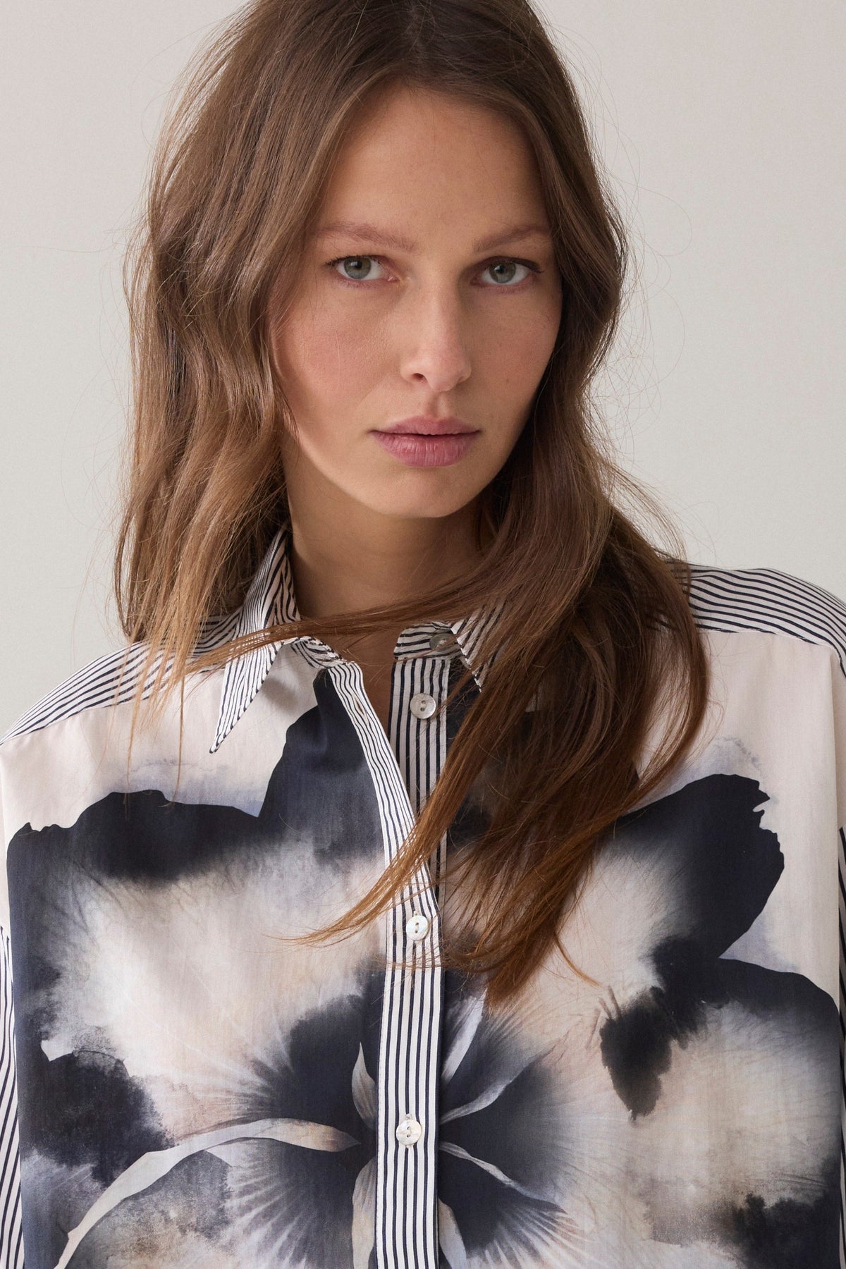 A woman  wearing a long-sleeve button-up shirt with thin vertical blue and white stripes, and statement floral design paired with dark pants, against a plain light beige background.
