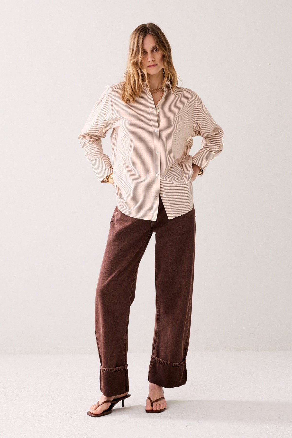 A woman modeling a light beige button-up shirt and dark brown wide-leg corduroy pants against a plain light gray studio background.