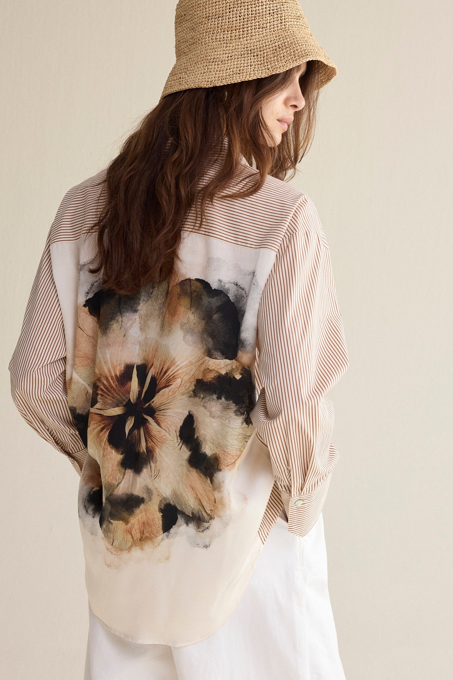 A woman wearing a beige straw bucket hat and a long-sleeve button-up shirt with a large abstract floral print in black, brown, and beige on the back, paired with white pants, photographed against a plain light beige background.