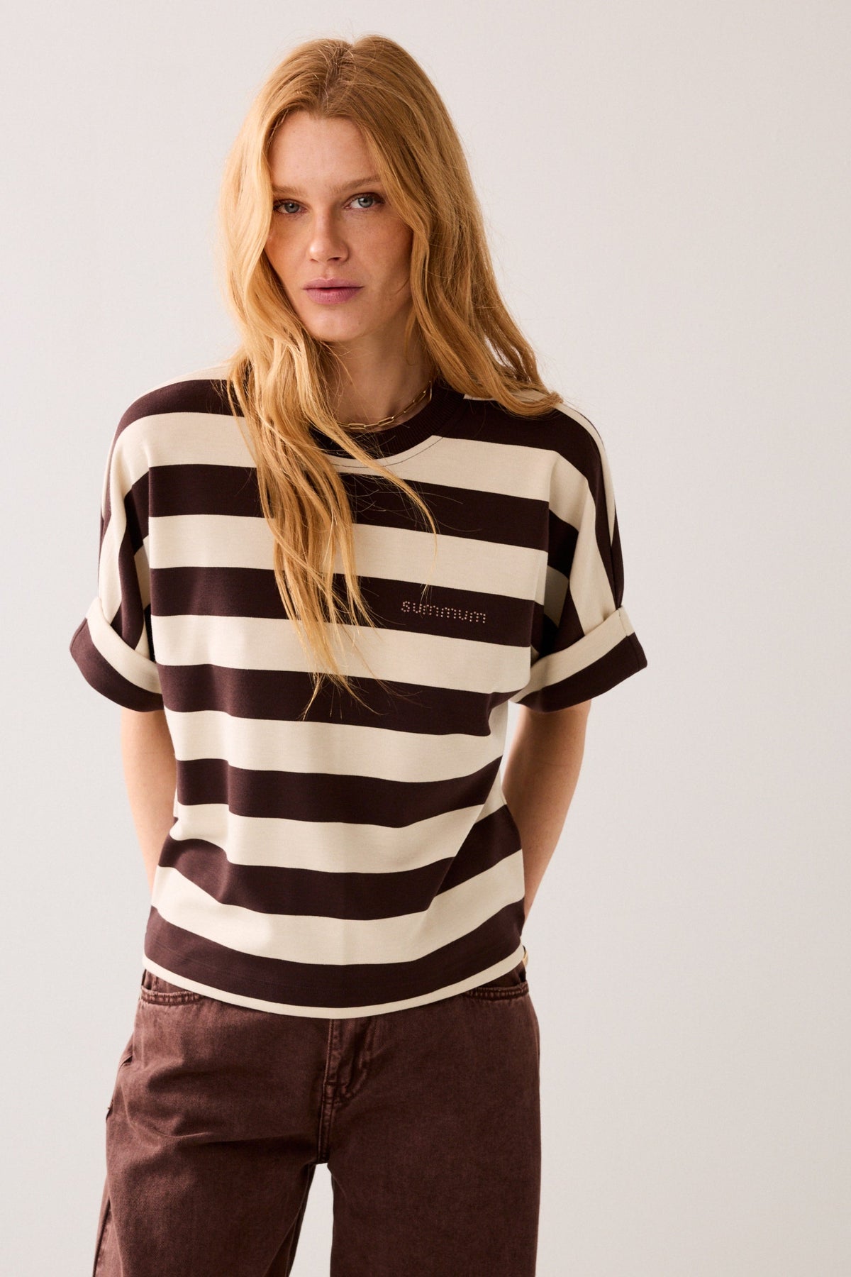 Woman wearing a short-sleeve crewneck t-shirt with horizontal dark brown and cream stripes, featuring a small embroidered 'summum' logo on the left chest, displayed on a plain background.