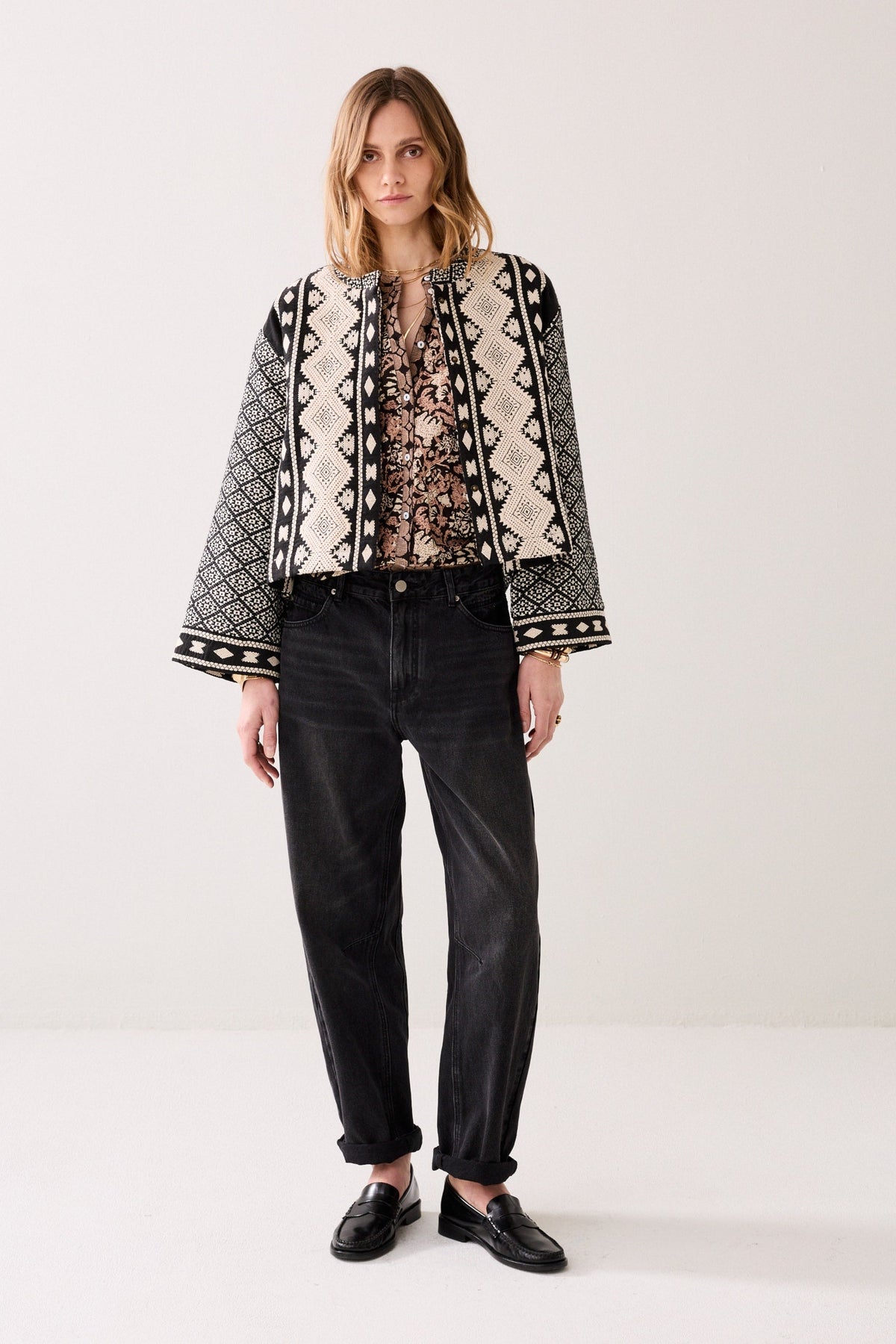 Woman wearing a patterned jacket, with a shirt underneath and grey jeans on a white background