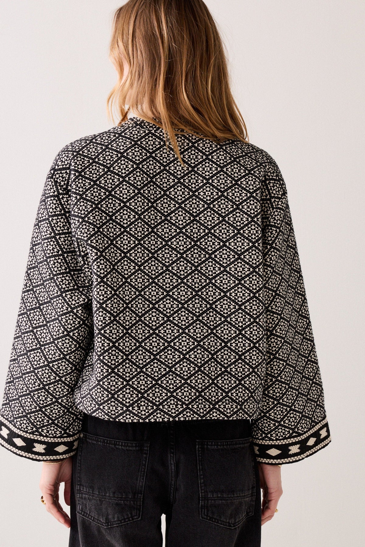 A woman viewed from the back wearing a black and off-white geometric patterned jacket with wide three-quarter sleeves and patterned cuffs, paired with black jeans, against a plain light background.