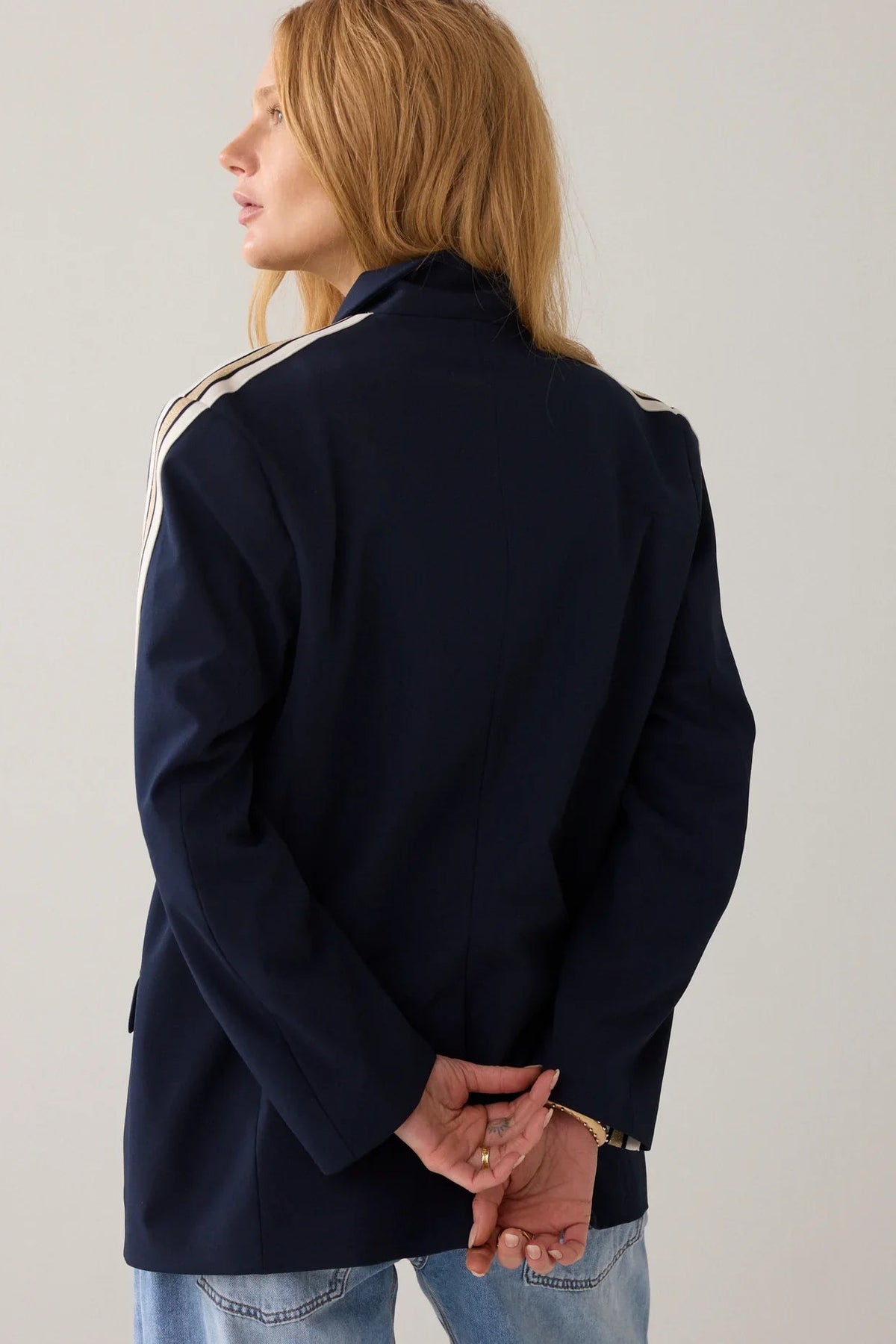 A women shown from the back wearing a  navy blue blazer with signature three-stripe detailing on the sleeves, featuring beige and white stripes, shown against a plain white background.