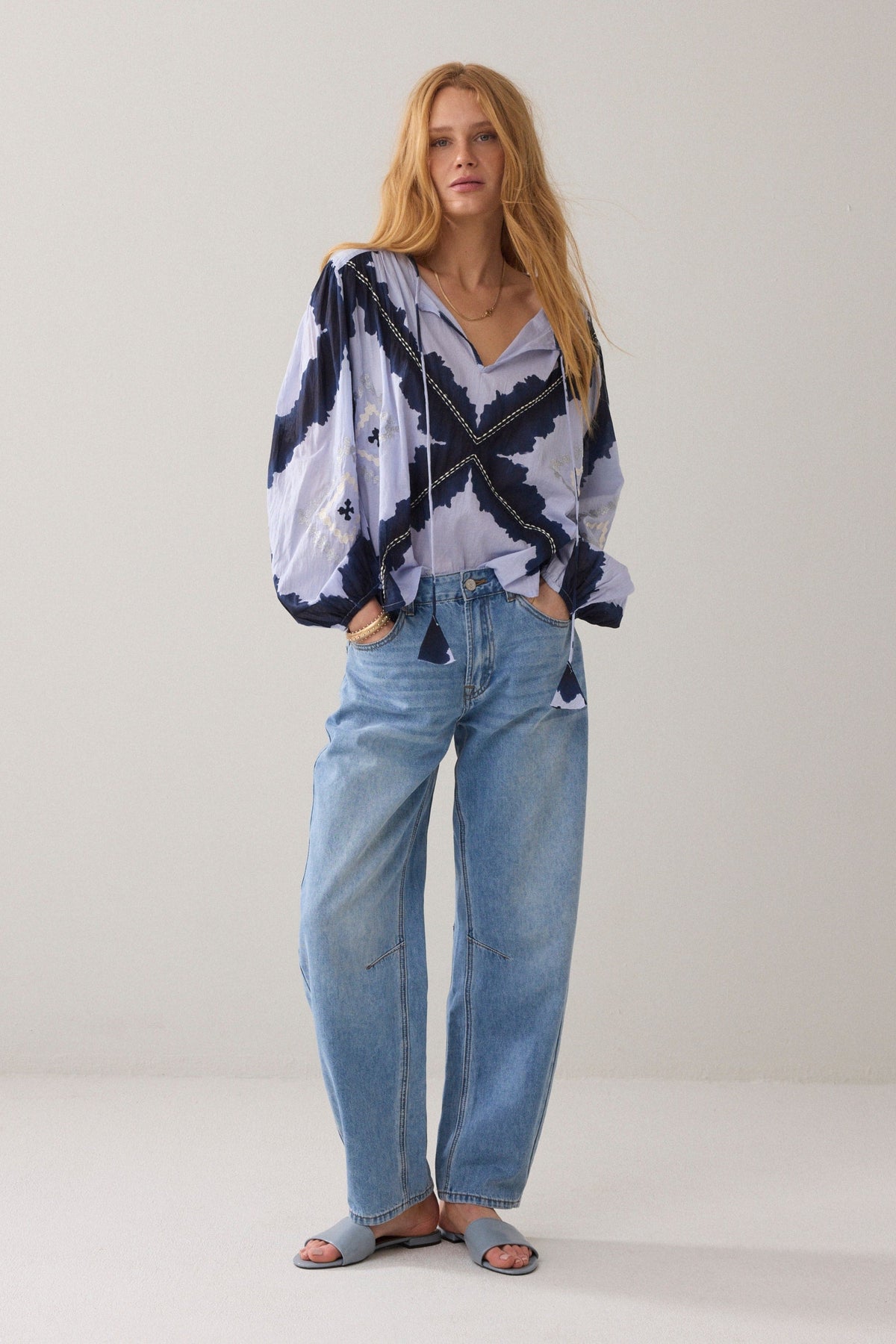 A woman  wearing a loose, long-sleeve blouse with a navy and pale lavender abstract pattern and decorative contrast stitching, paired with light blue jeans against a plain light background.