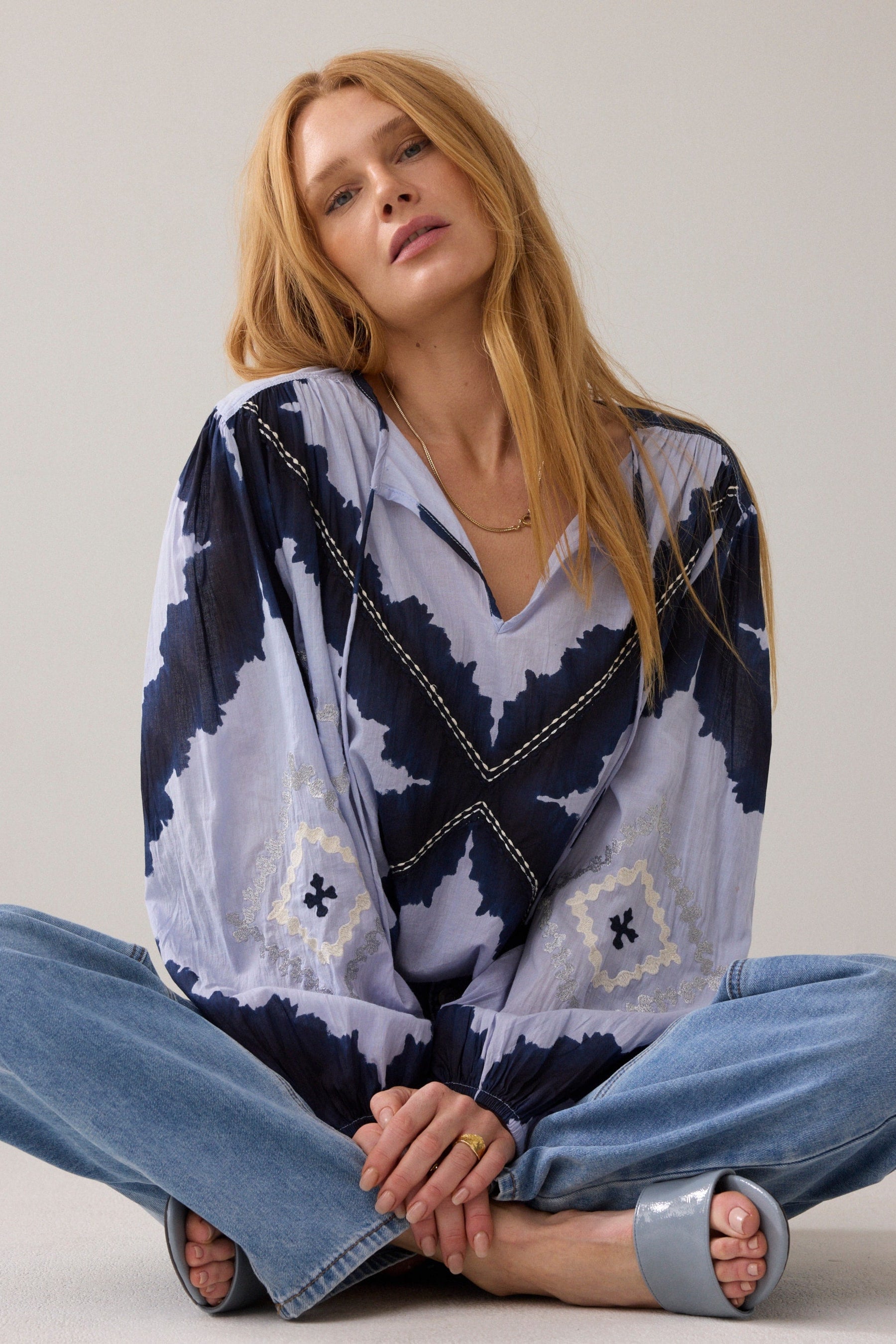 A woman sitting cross legged wearing a loose, long-sleeve blouse with a navy and pale lavender abstract pattern and decorative contrast stitching, paired with light blue jeans against a plain light background.