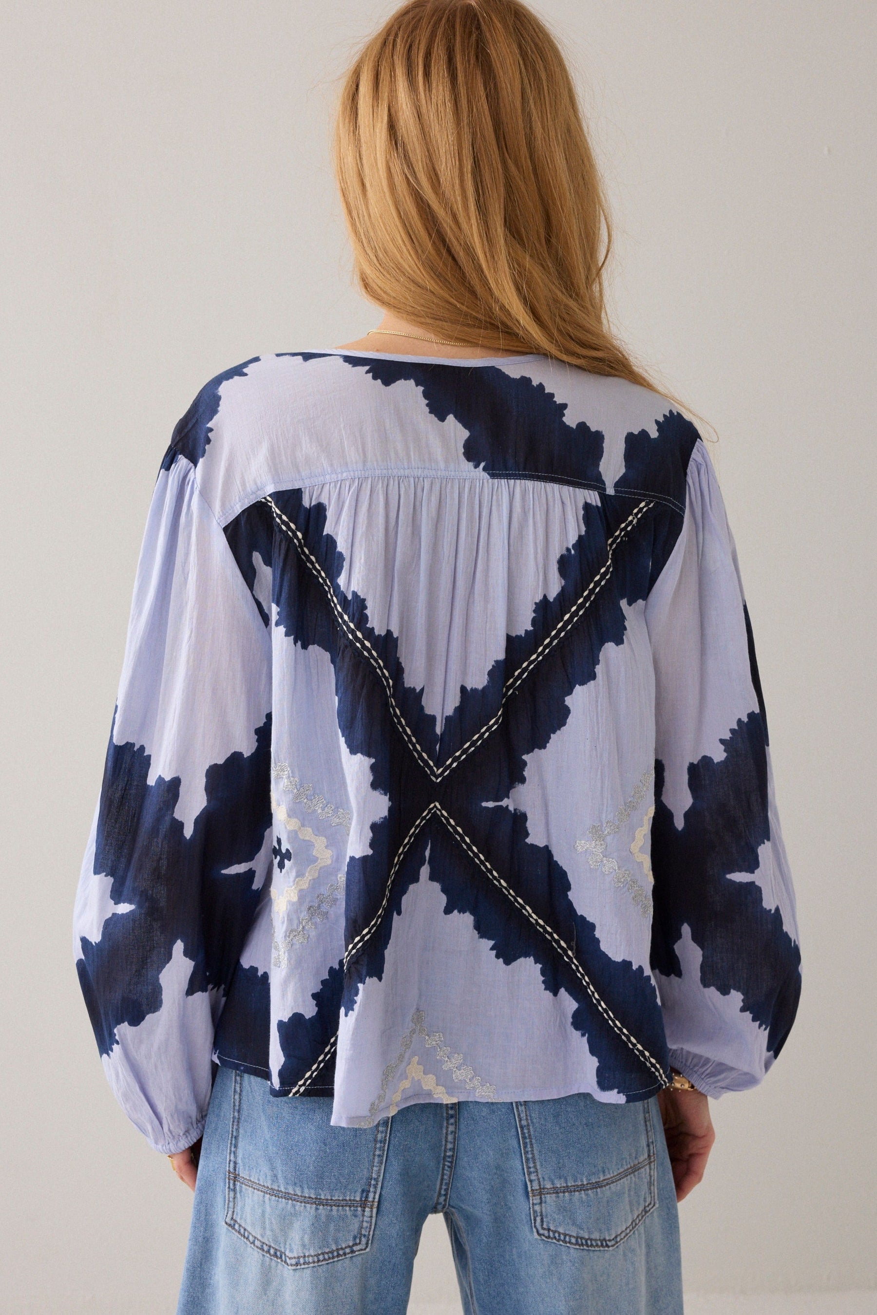 A woman viewed from behind wearing a loose, long-sleeve blouse with a navy and pale lavender abstract pattern and decorative contrast stitching, paired with light blue jeans against a plain light background.