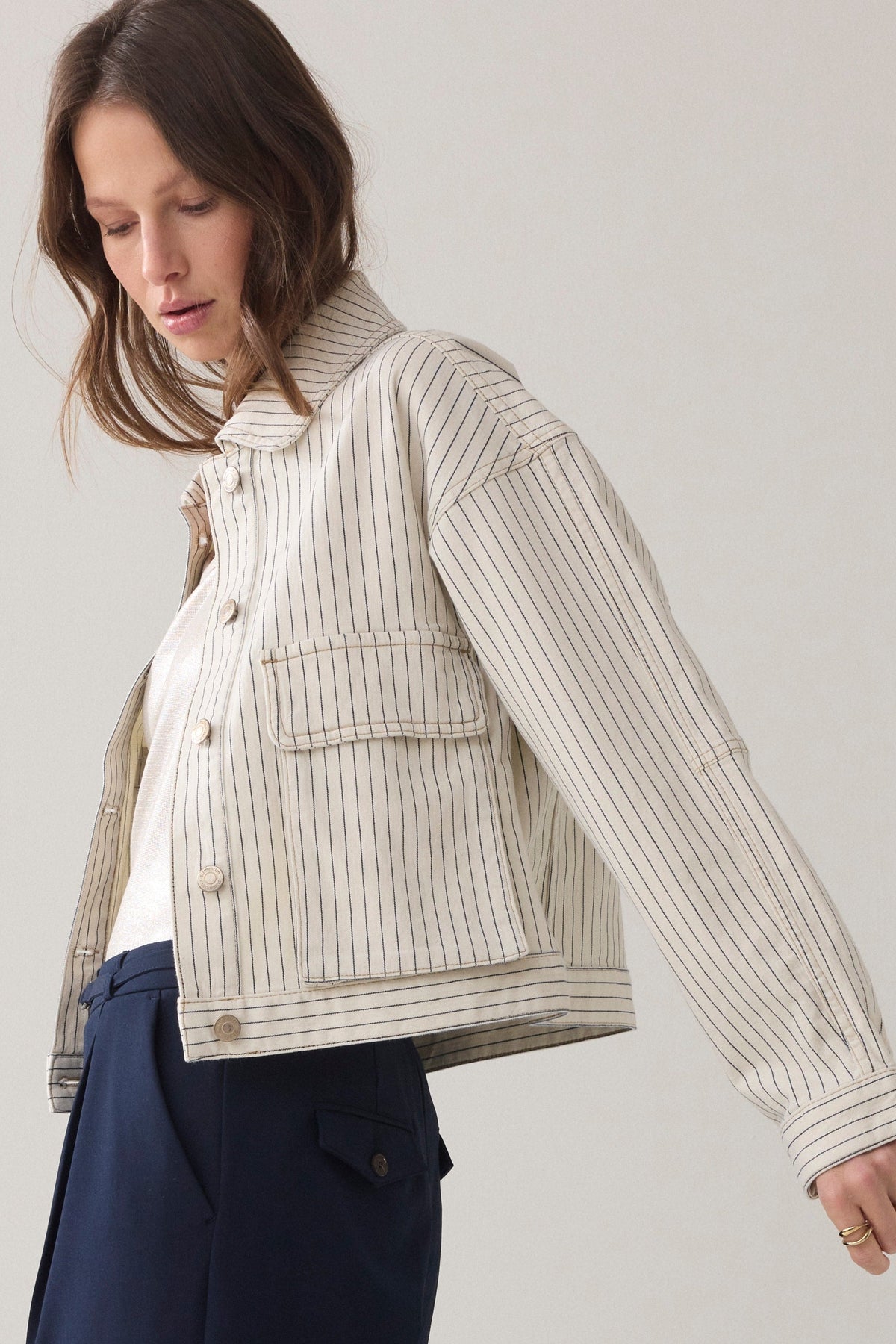 A women wearing cropped denim jacket wth an ivory base and navy herringbone vertical stripe detail  and navy wide leg paints and light coloured sneakers on a plain background.  